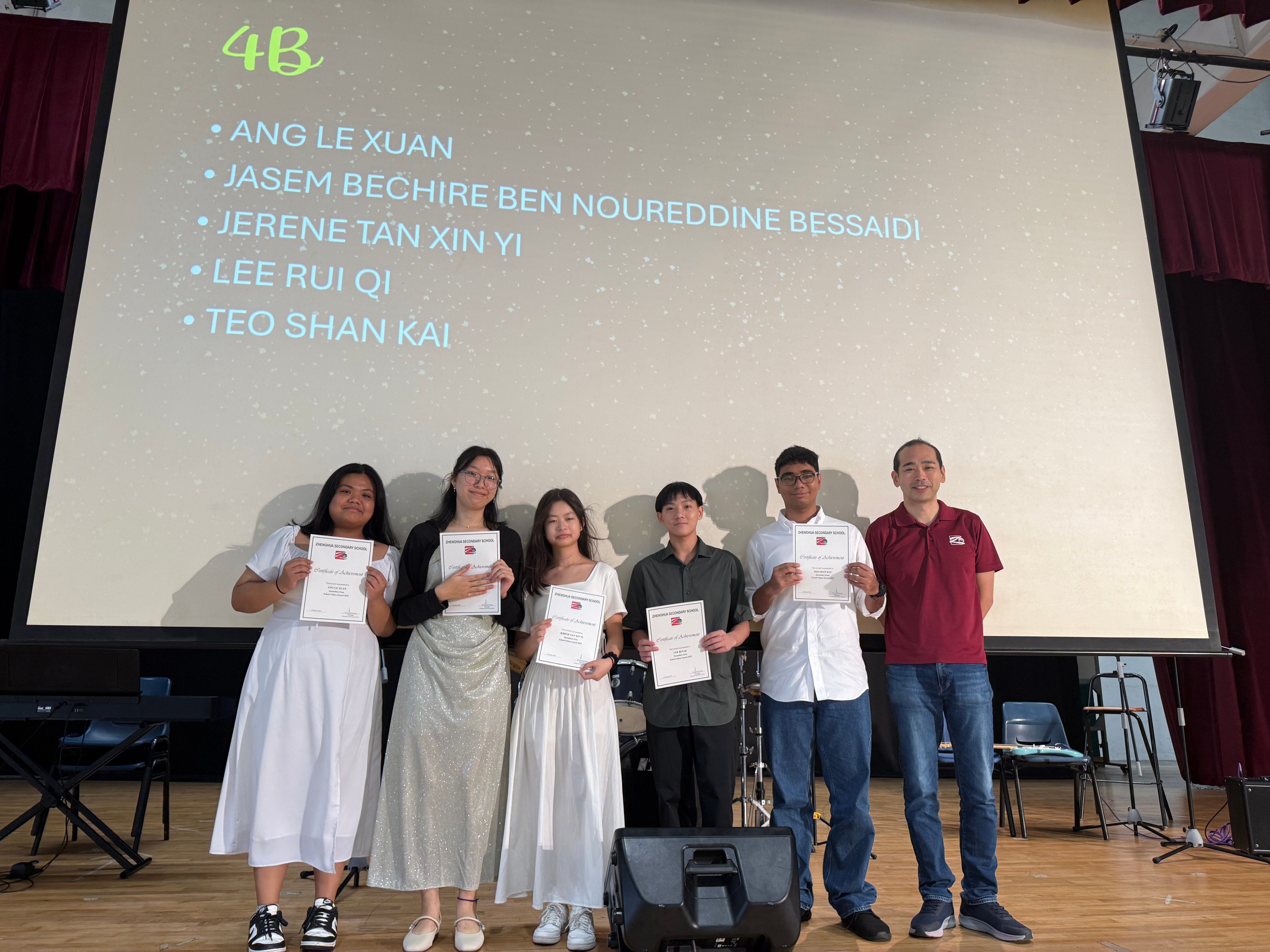 Five students and a teacher stand on a stage holding certificates, with a projection screen showing class 4B names behind them.