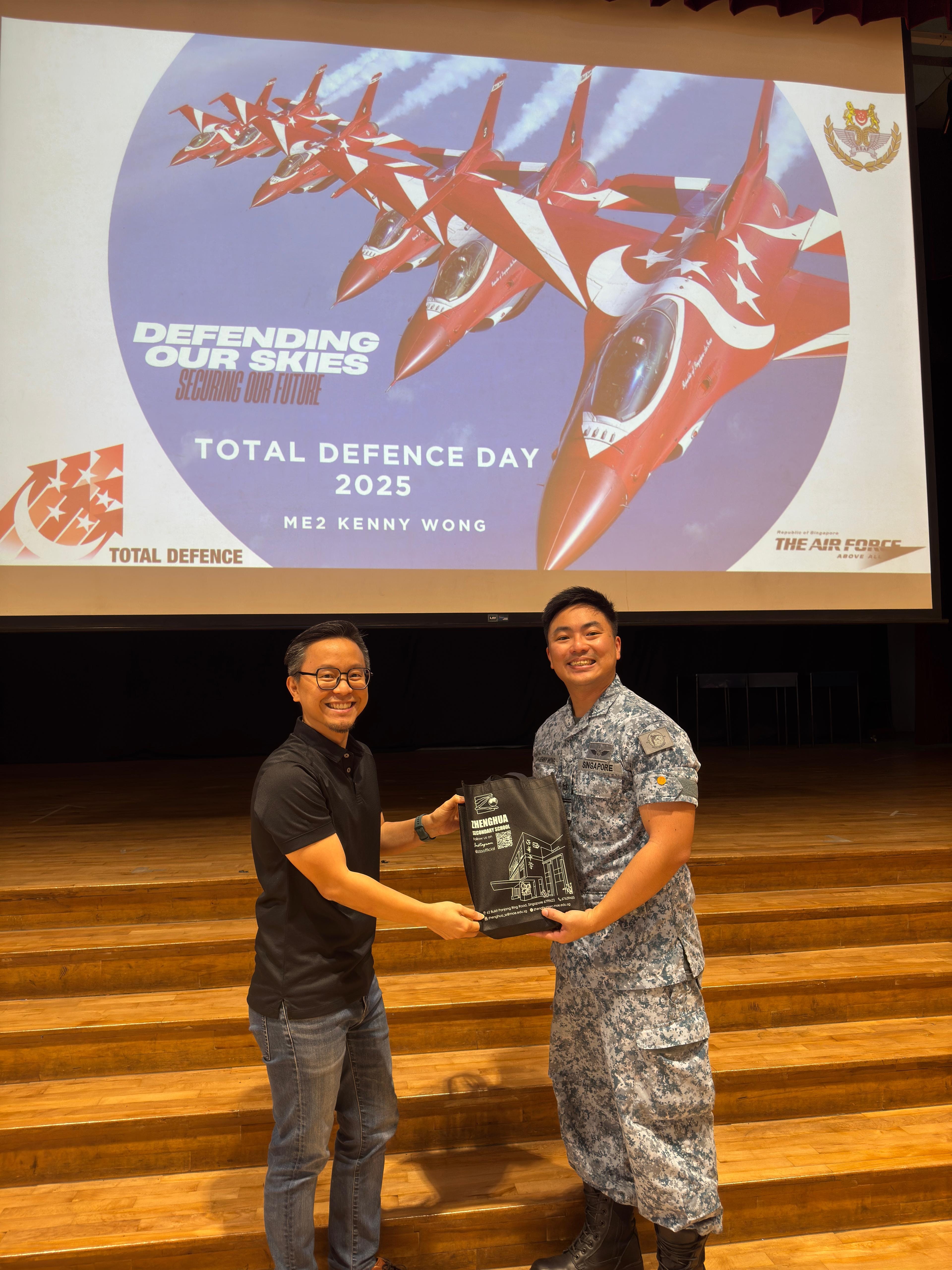 Two men smile while holding a bag in front of a Total Defence Day 2025 presentation screen showing red jets.