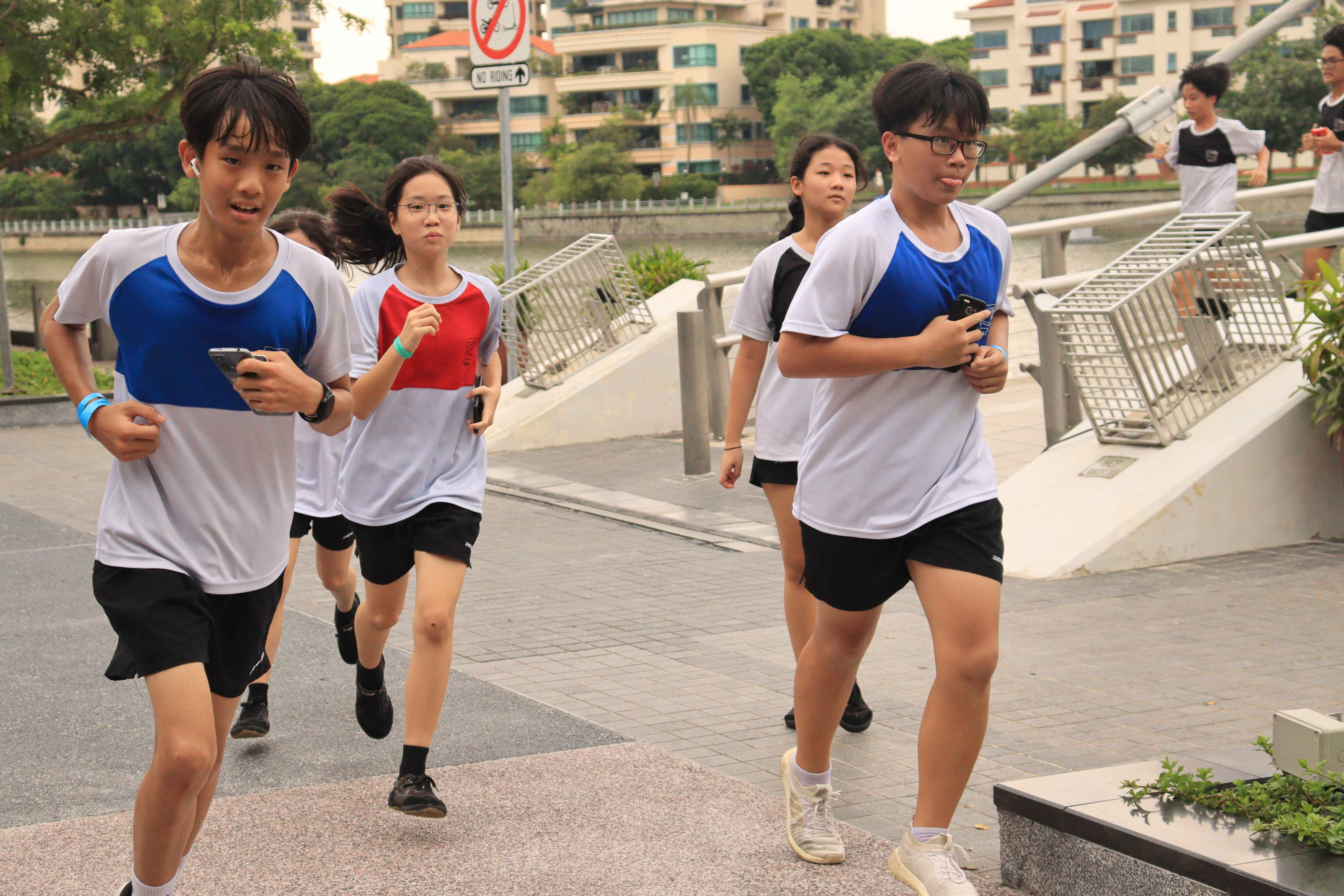 A group of teenagers in white and blue sports uniforms jogging along an outdoor waterfront path.
