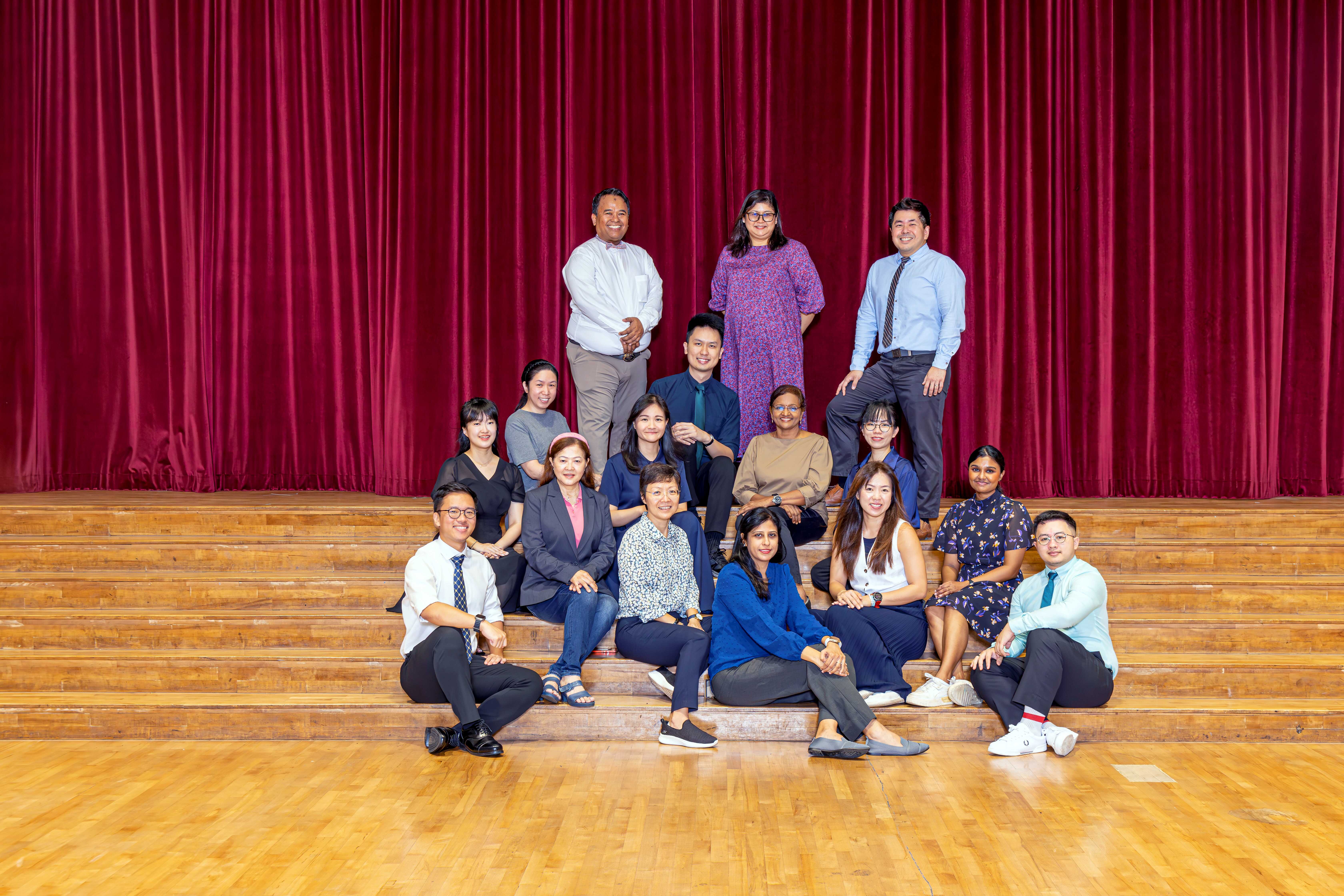 A group of about 15 people posing on wooden stage steps in front of a red curtain, smiling at the camera.