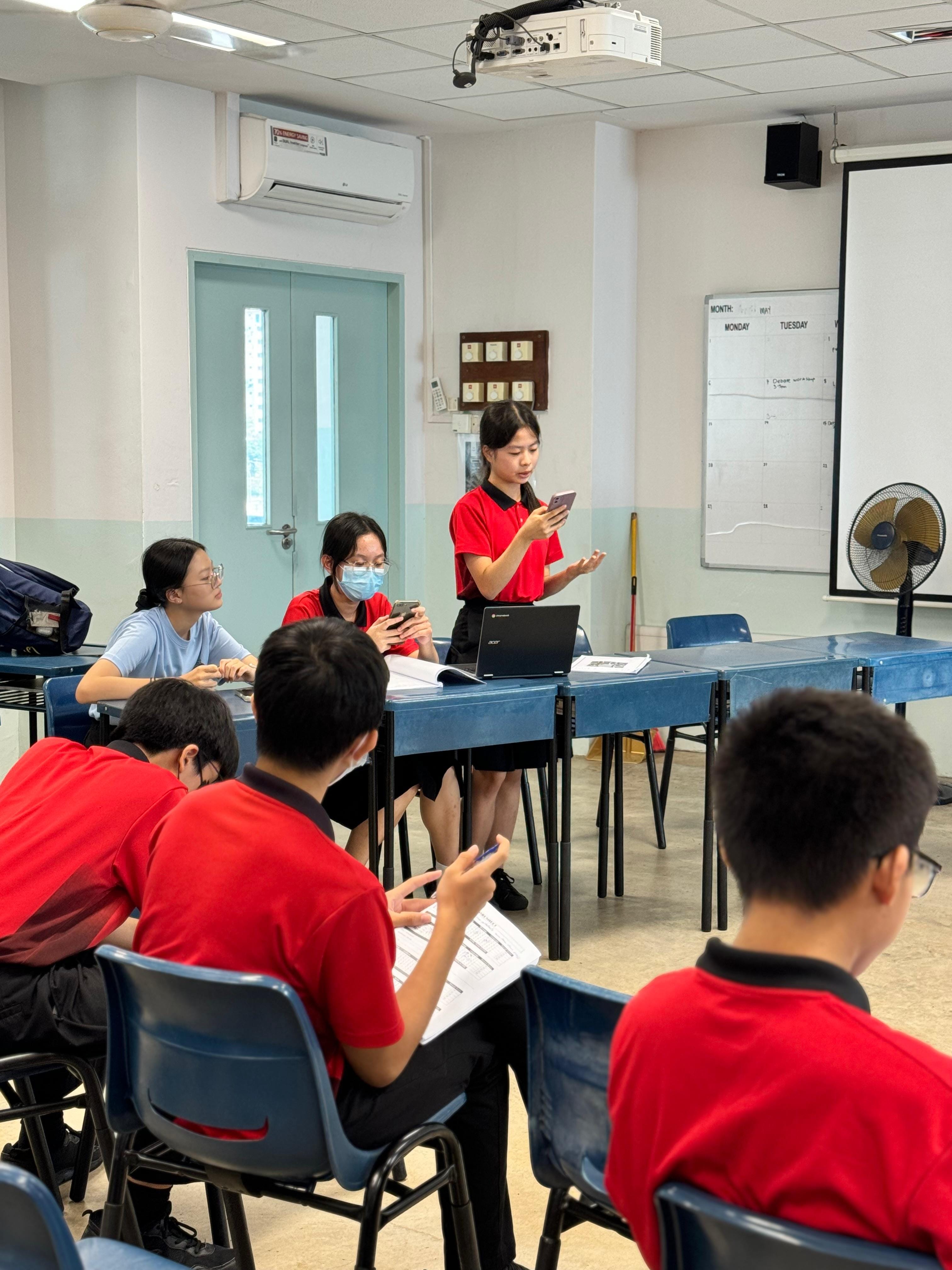 Students in red uniforms sit at blue desks in a classroom while one student stands holding a phone.