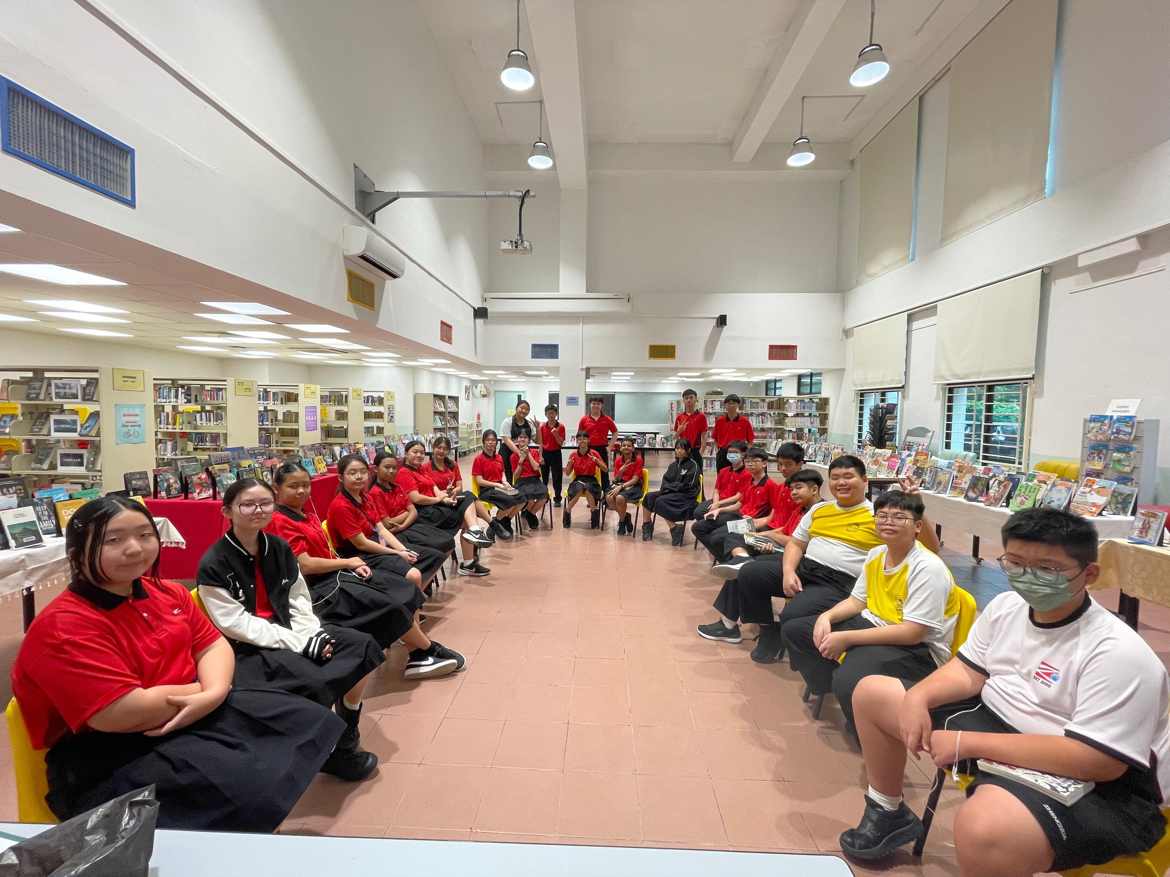 A large group of students in red and black uniforms sit in a circle inside a school library with bookshelves around them.