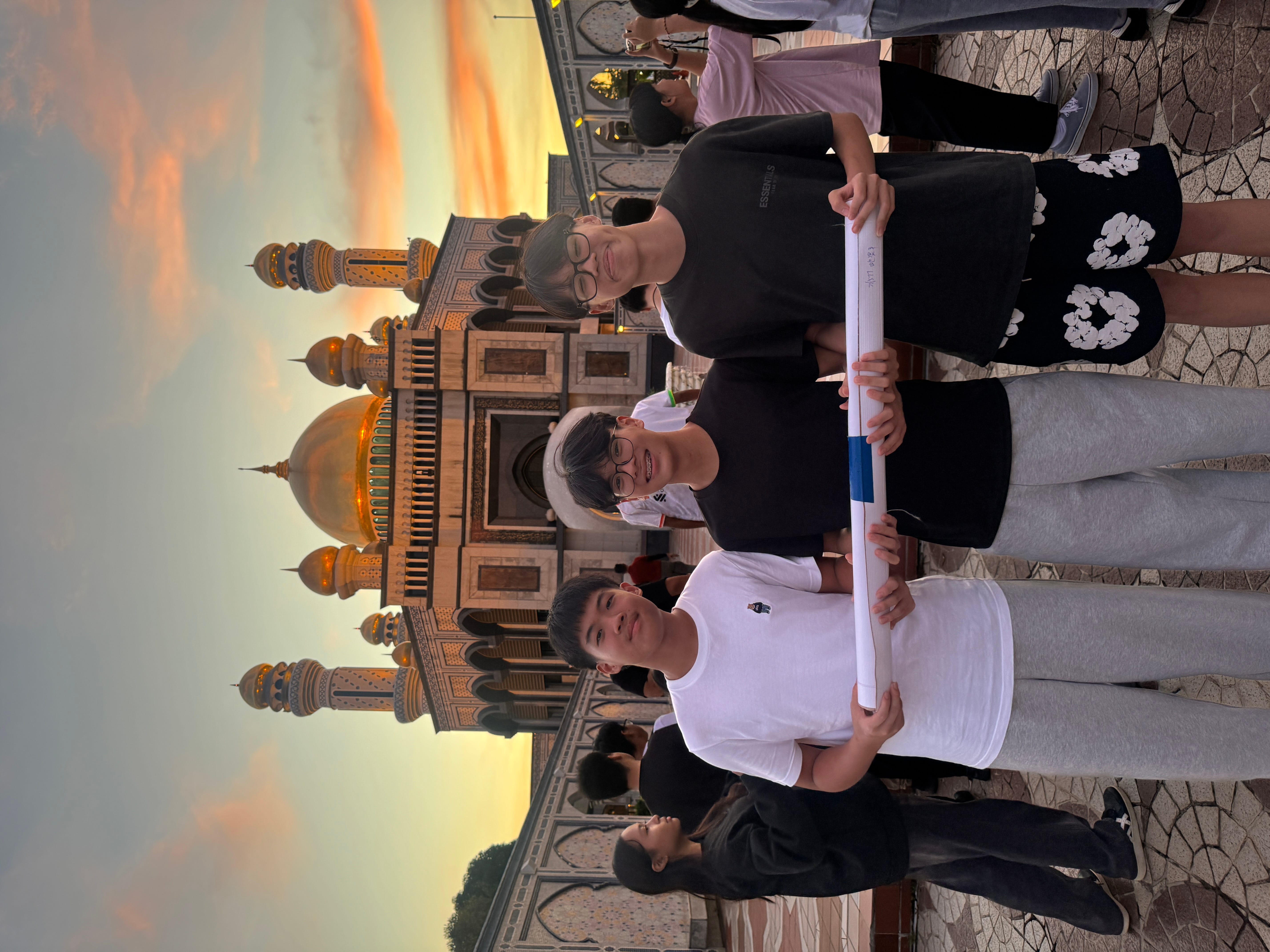 Three boys pose holding a rolled paper in front of a domed mosque at sunset, with people behind.