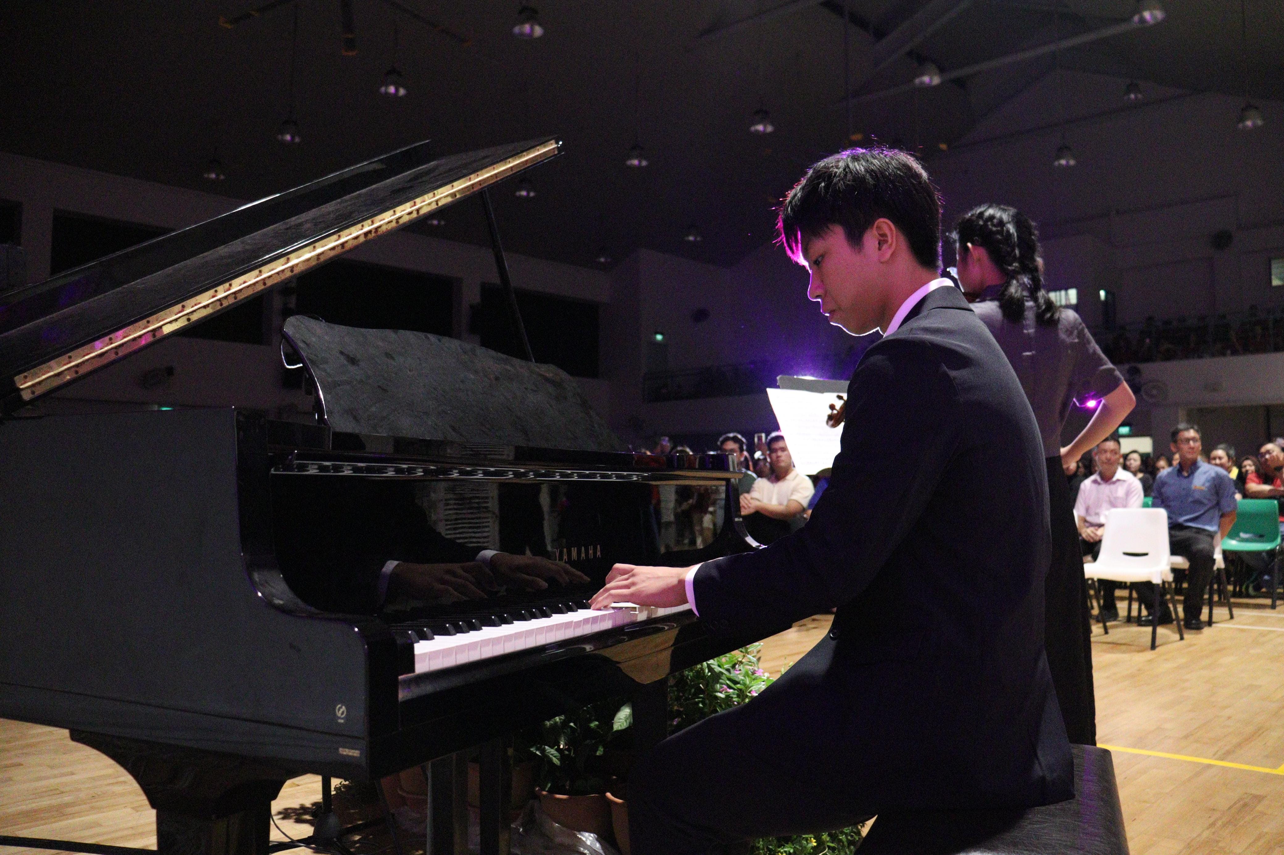 Young man in a black suit plays a Yamaha grand piano on a dimly lit stage before a seated audience.