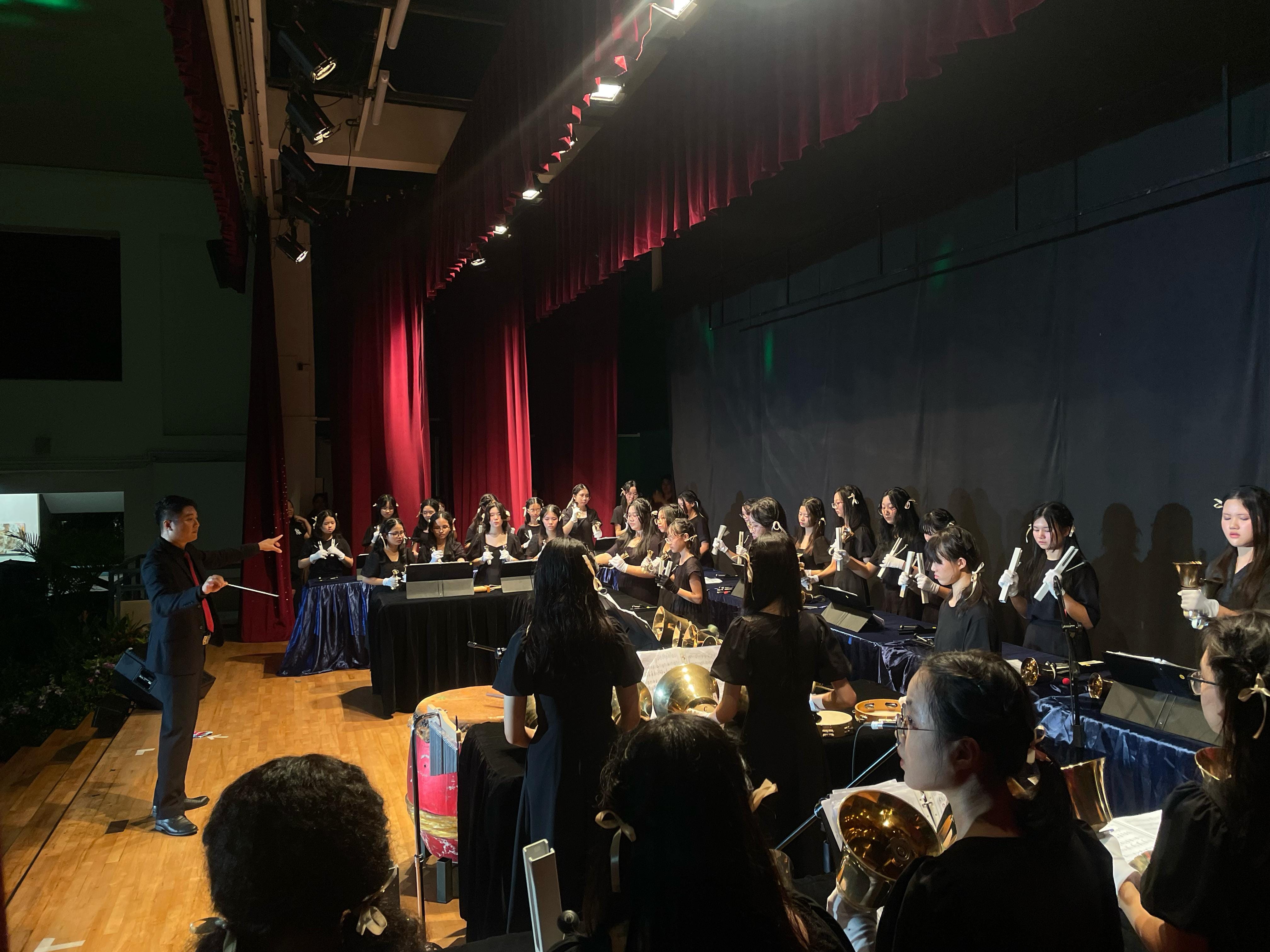A conductor in a black suit leads a youth ensemble in black outfits performing with handbells on a stage with red curtains.