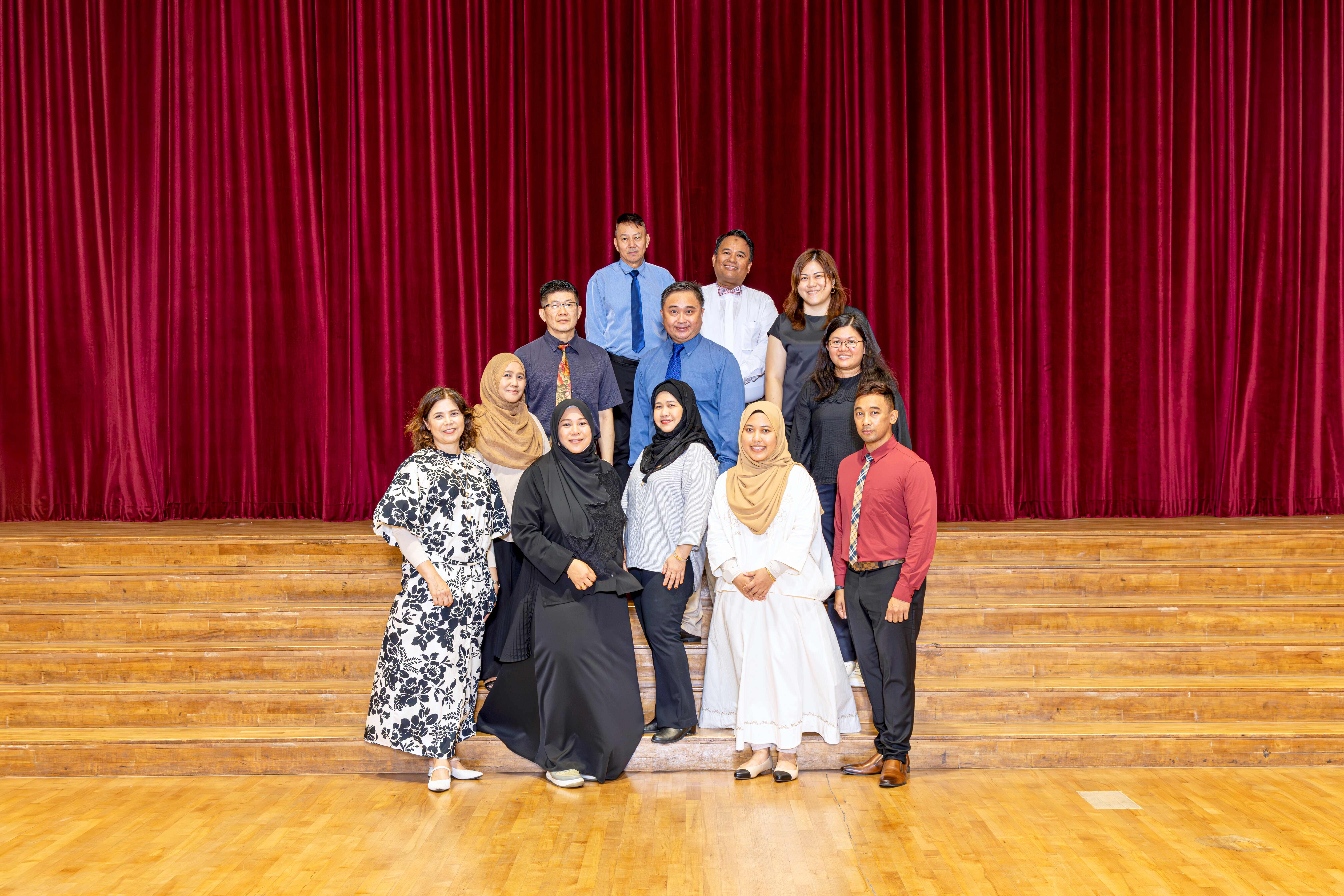 A group of eleven people posing in rows on wooden stage steps before a red velvet curtain.