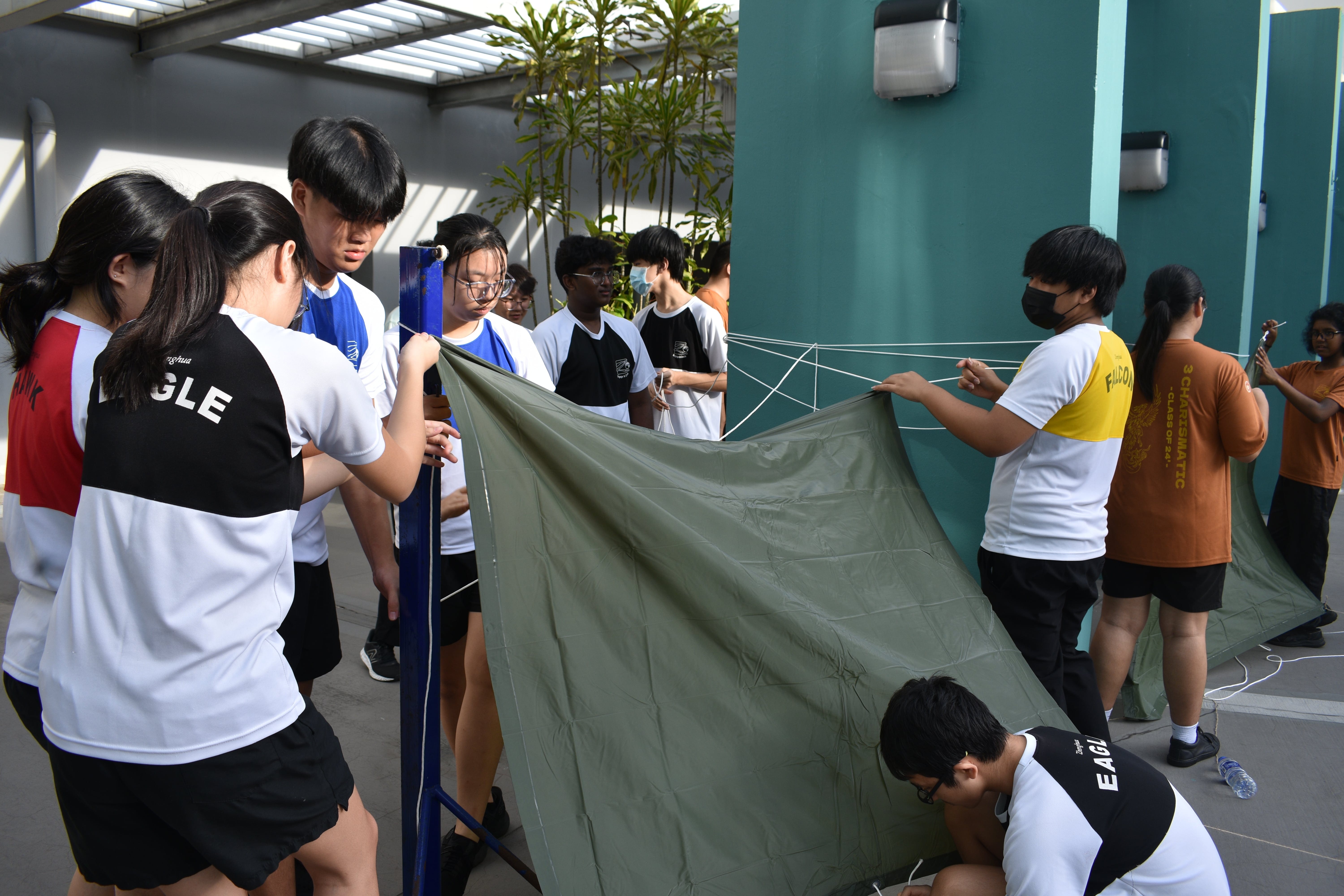 A group of students in school uniforms working together to set up a green canvas tent outdoors.