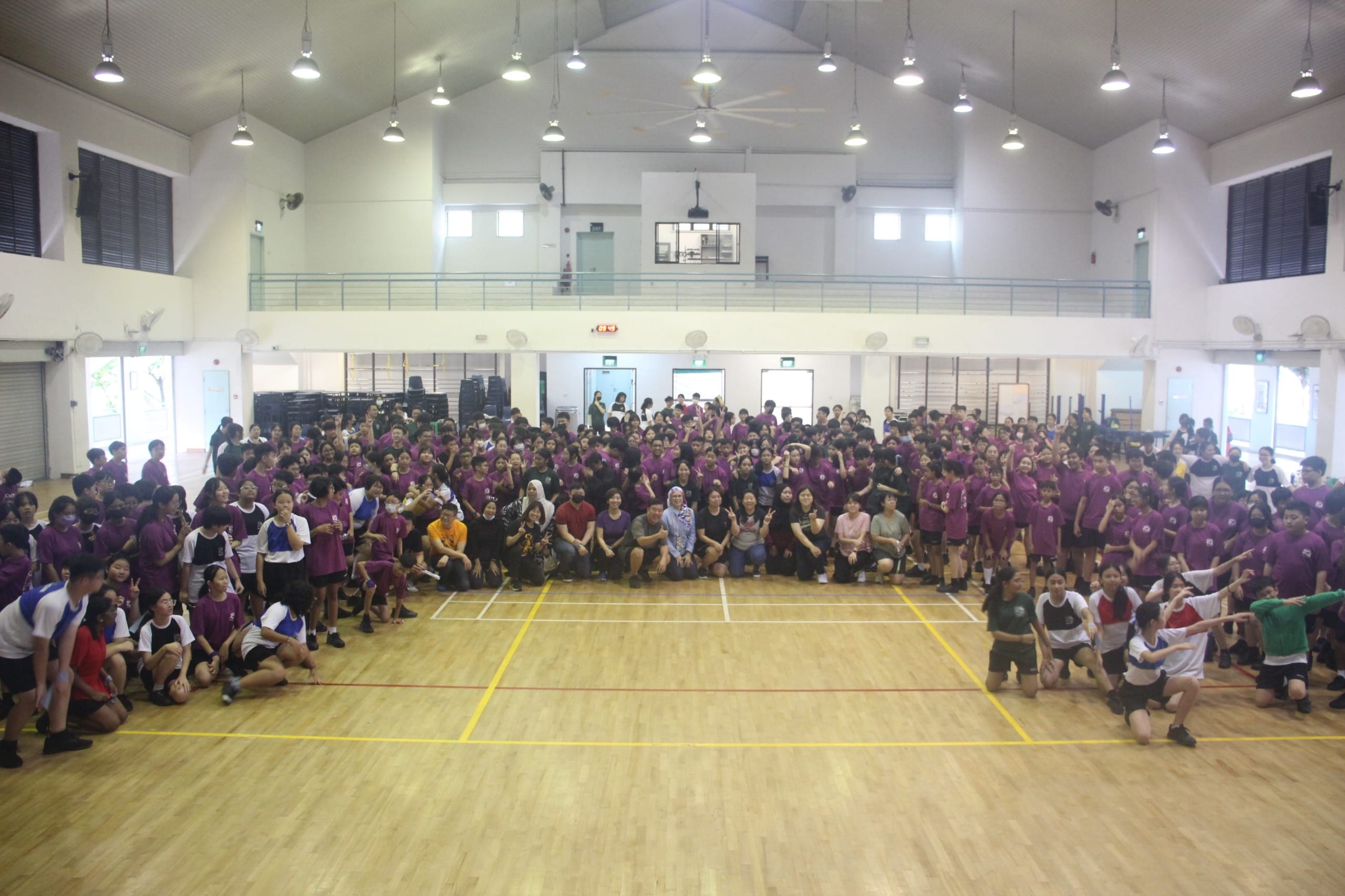 Large group of students mostly wearing purple shirts gathered for a group photo inside a school gymnasium.