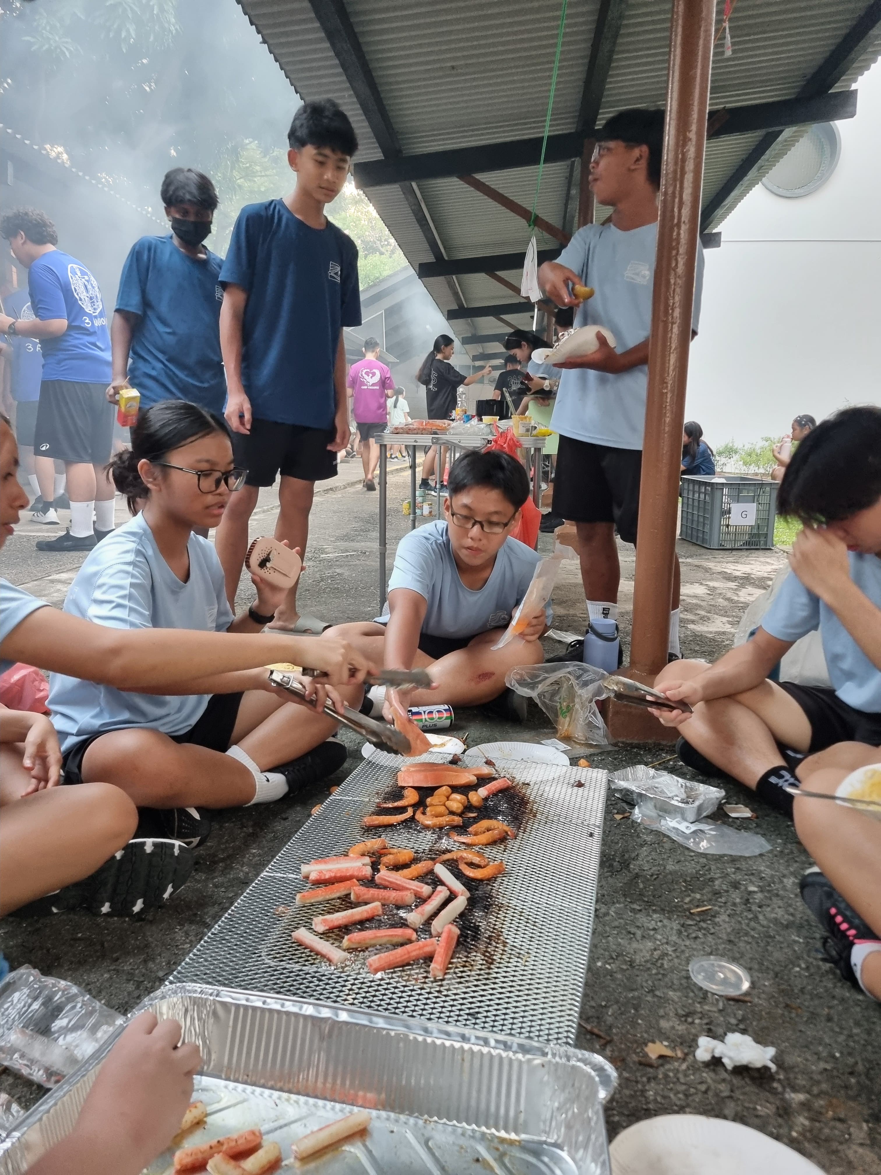 Teenagers in blue shirts sit on the ground grilling sausages and seafood on a flat charcoal grill under a smoky shelter.