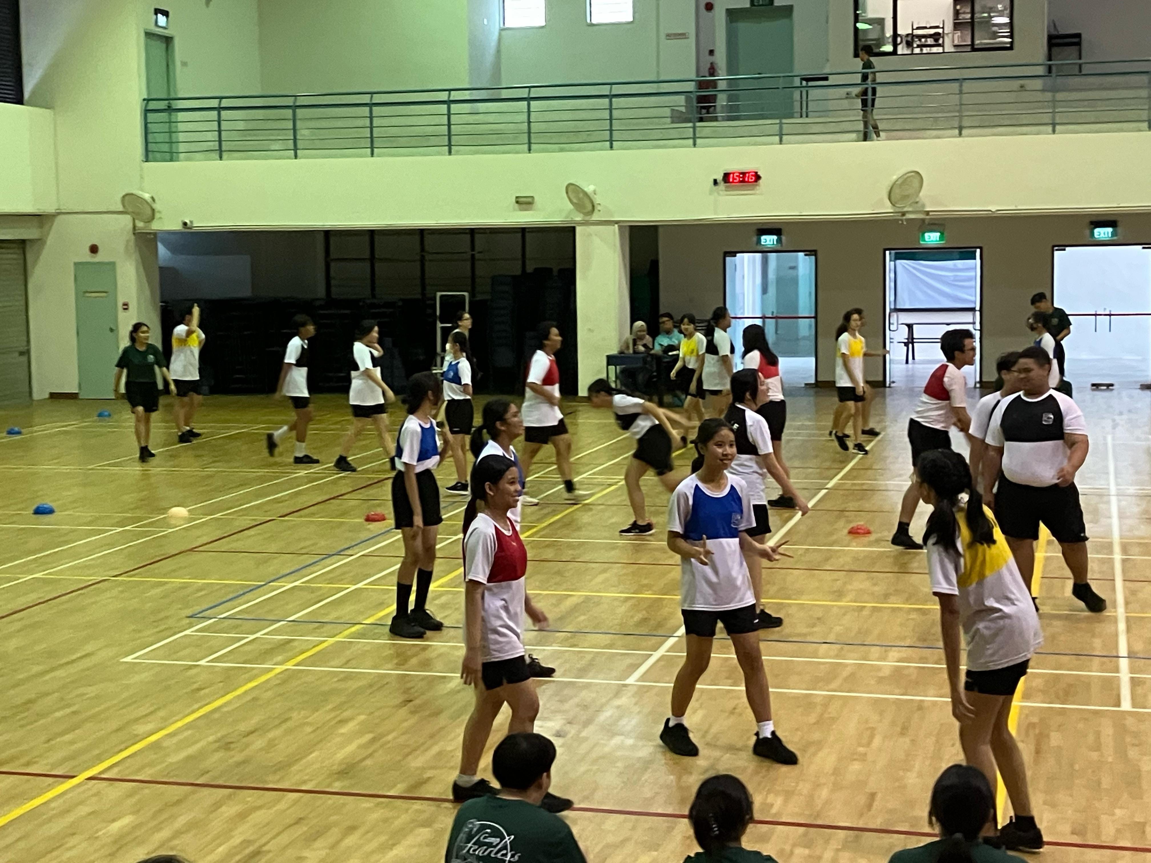 Students in white and colorful bibs play a sport on an indoor wooden gymnasium court.