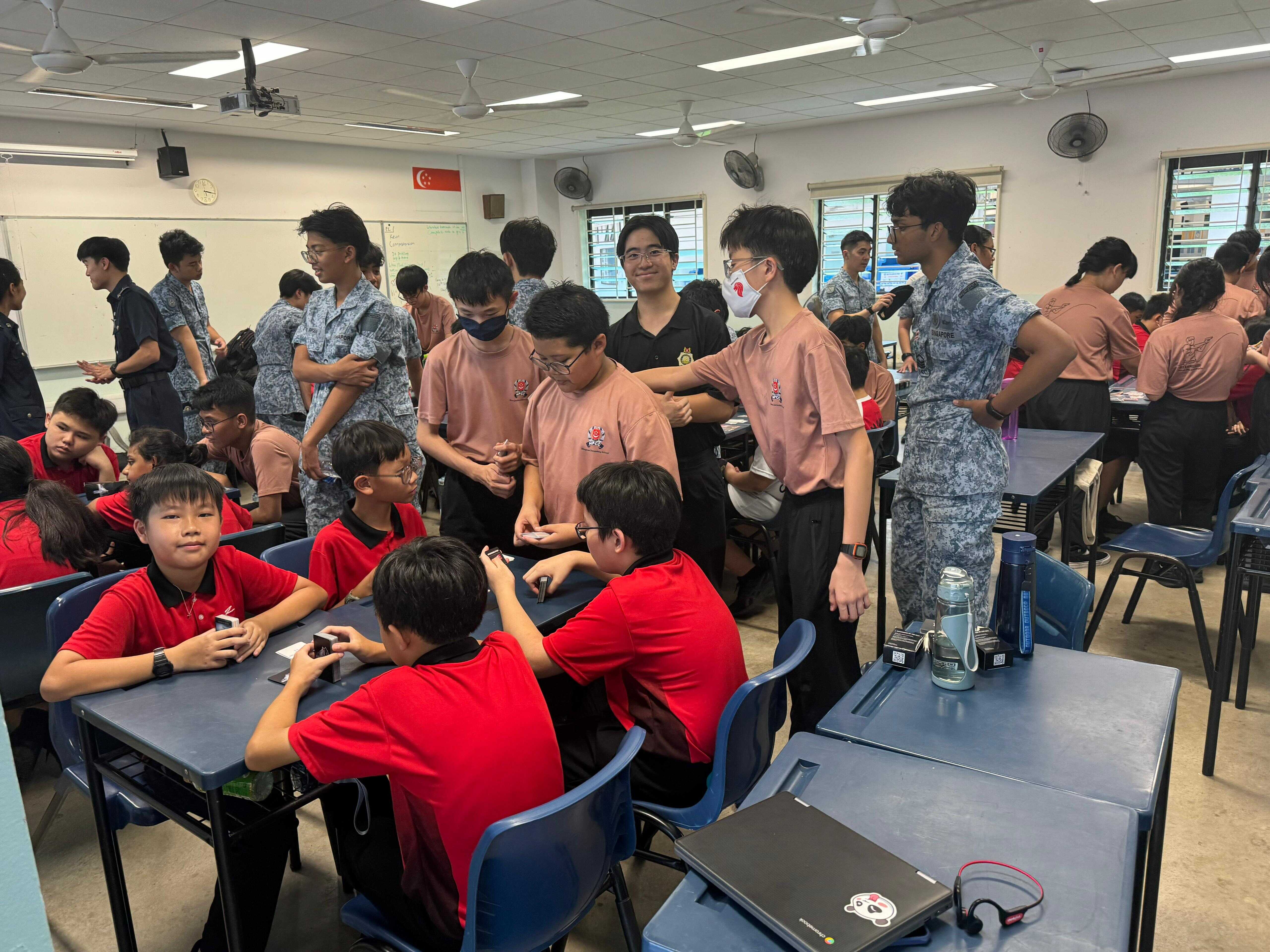 Students in red, pink, and camouflage uniforms gather in a busy classroom with desks, fans, and a Singapore flag.