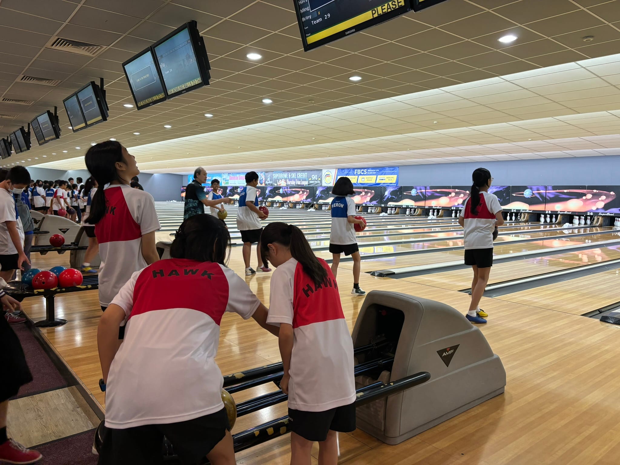 Youth bowlers in red and white Hawk jerseys compete on multiple lanes in a busy bowling alley.