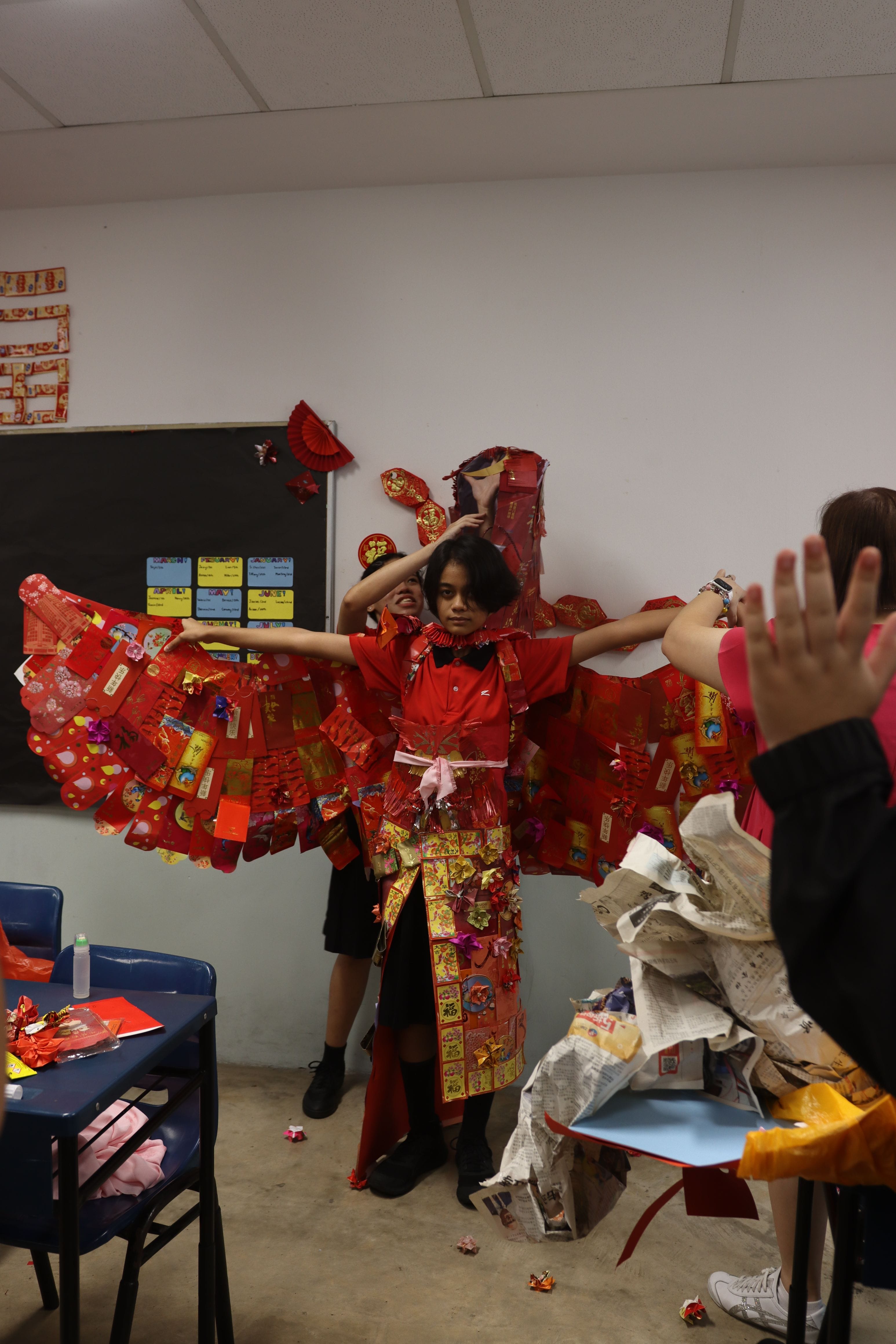 A child in a classroom wears an elaborate costume made of red Chinese New Year envelopes with arms outstretched.