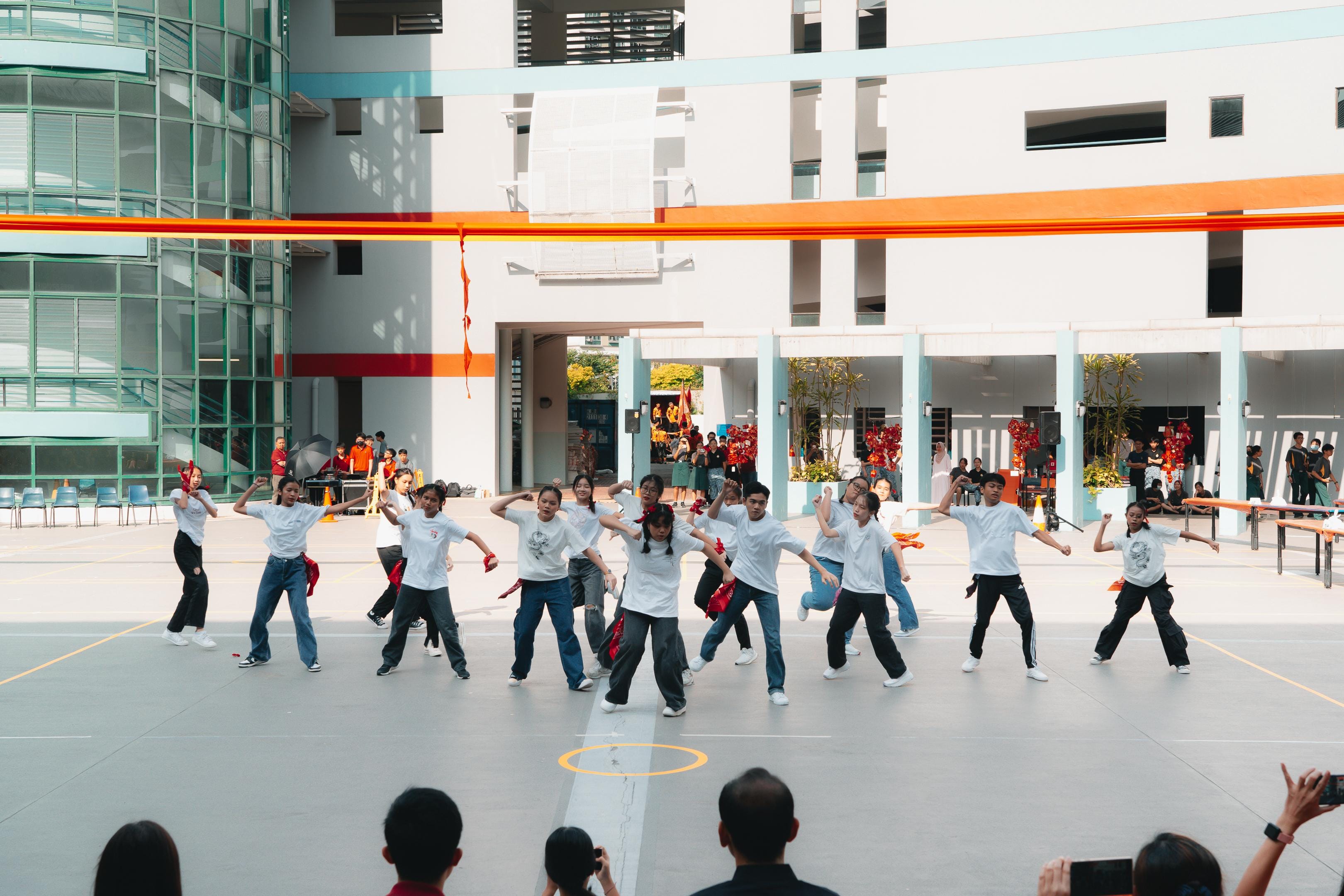A group of students in white shirts perform a dance routine outdoors in a school courtyard with orange banners overhead.