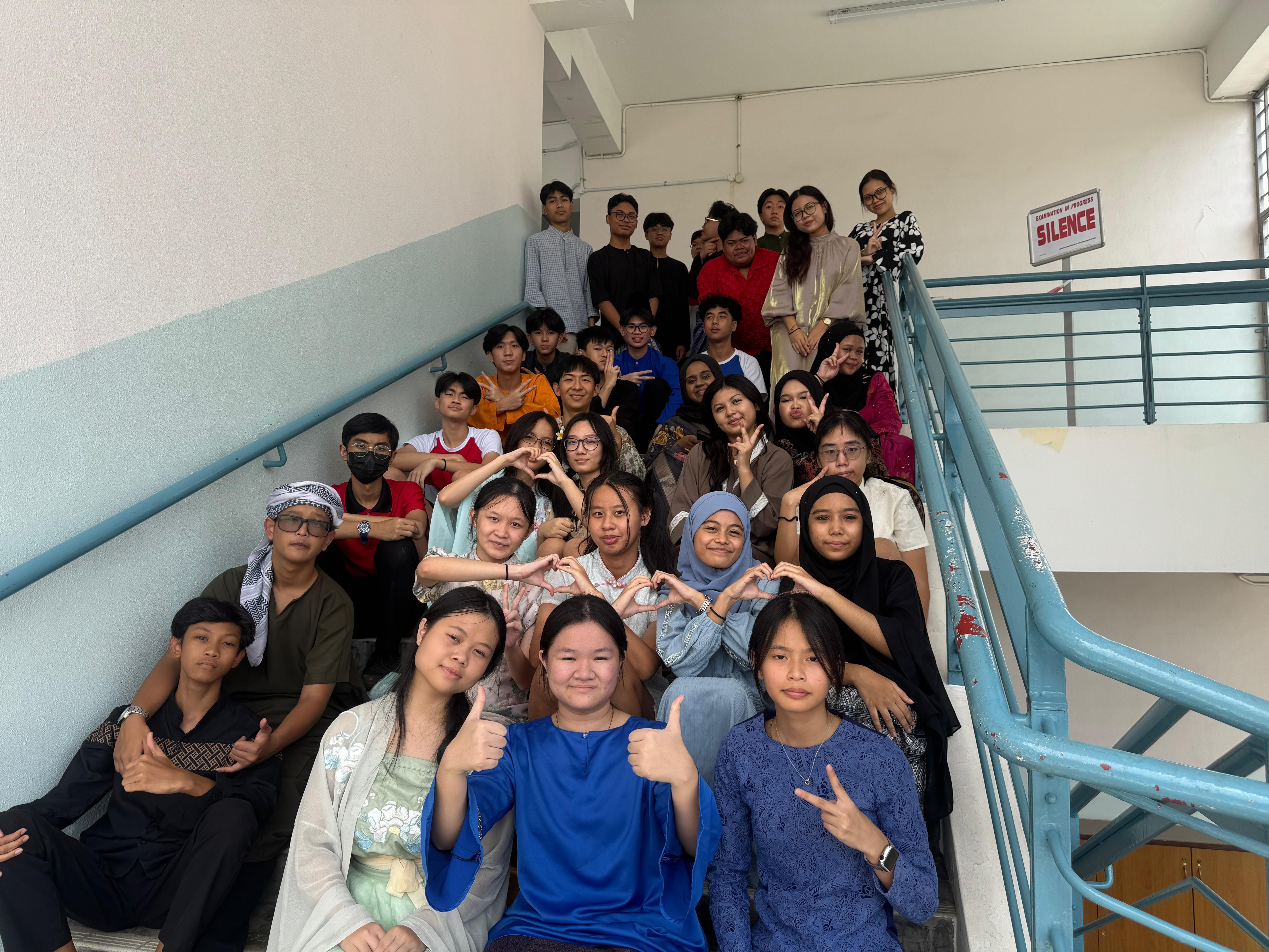A large group of students posing on indoor stairs, making heart and peace signs, with a Silence sign on the wall.
