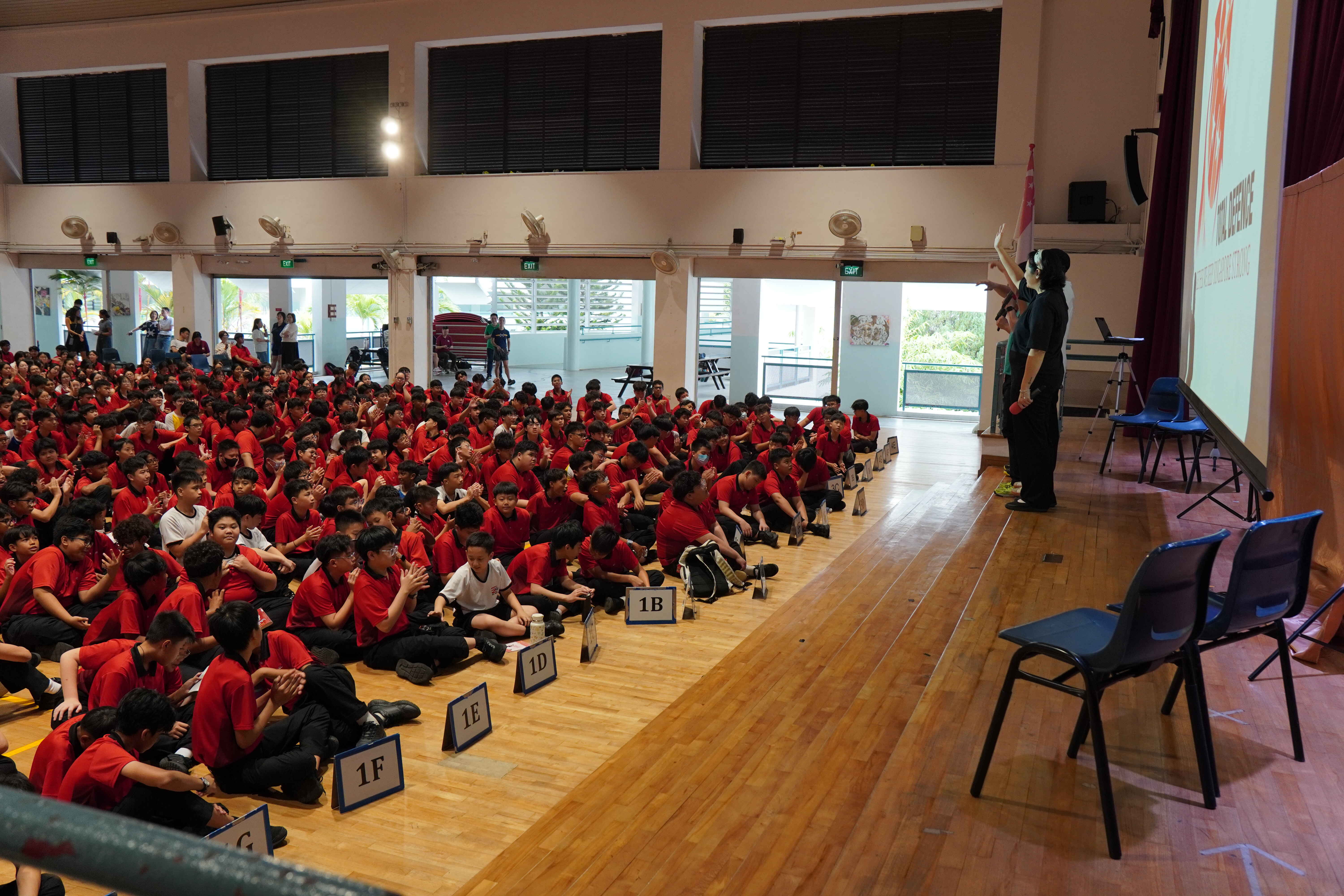 Large group of students in red shirts sitting on a gymnasium floor watching a presenter near a projected screen.