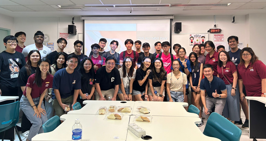 Large group of students posing in a classroom at a ZSS Alumni Day event, with snacks on tables.