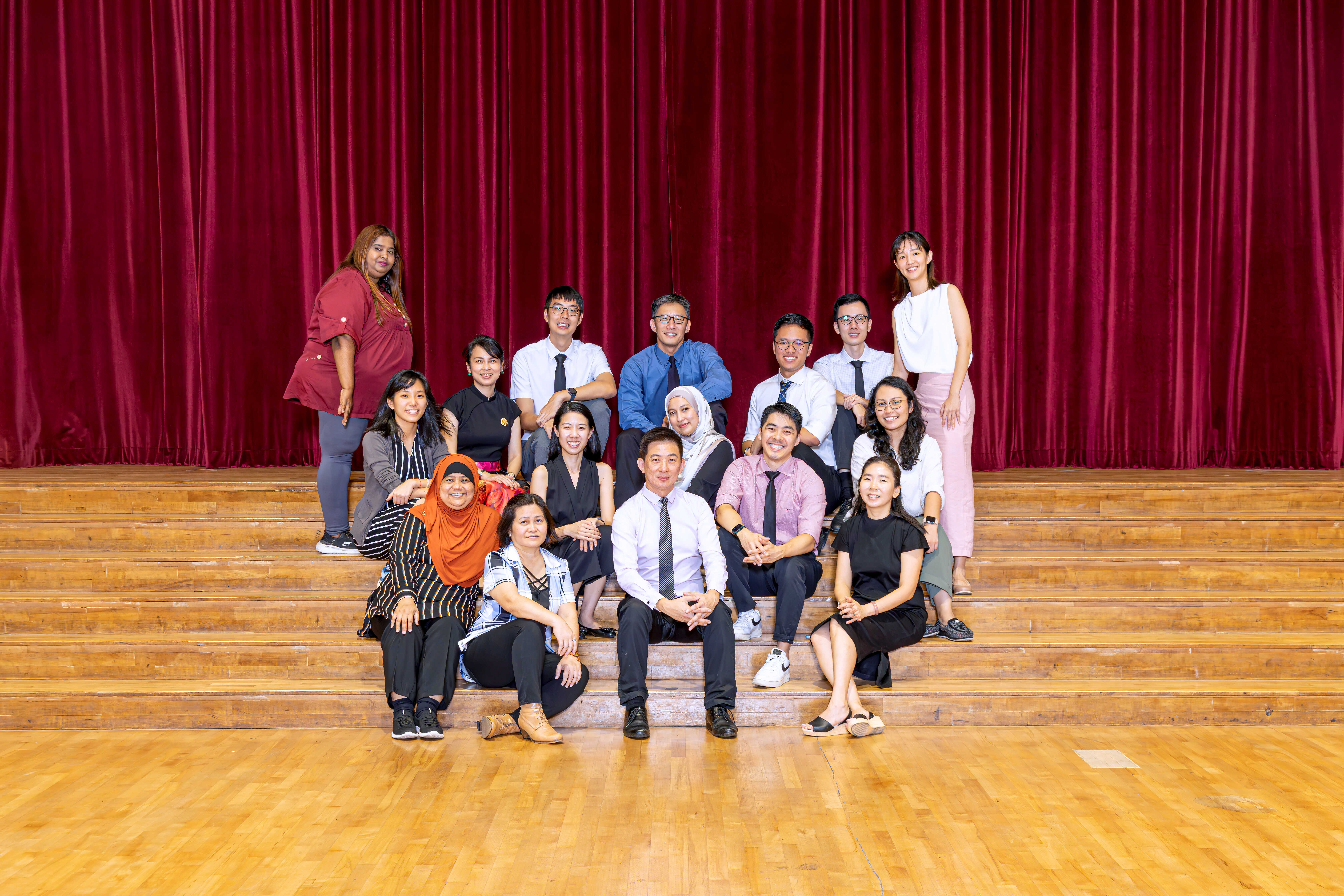 A group of about 15 people posing on wooden stage steps in front of a red curtain, smiling at the camera.