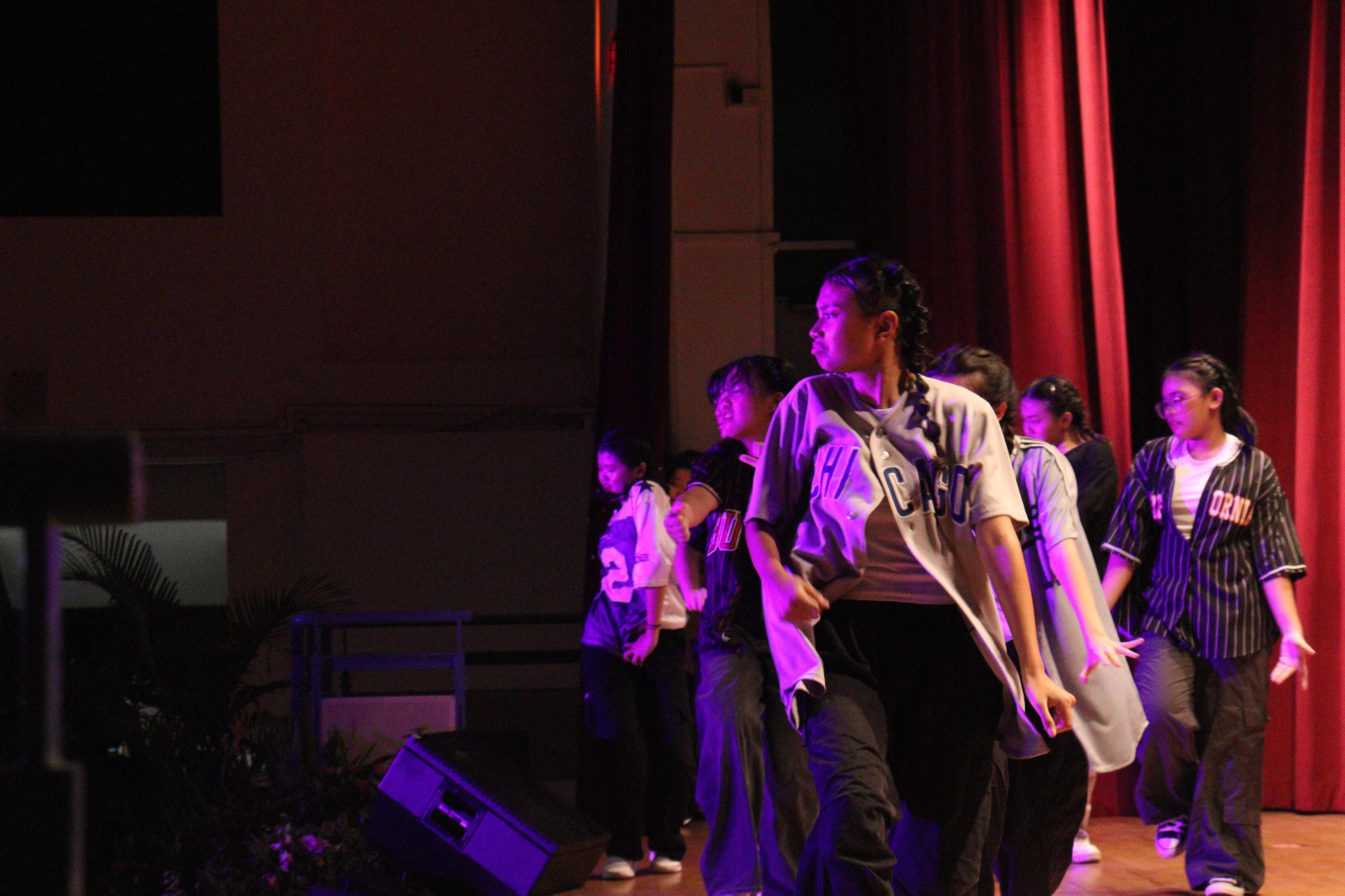 A group of young dancers in baseball jerseys perform on a dimly lit stage with red curtains.