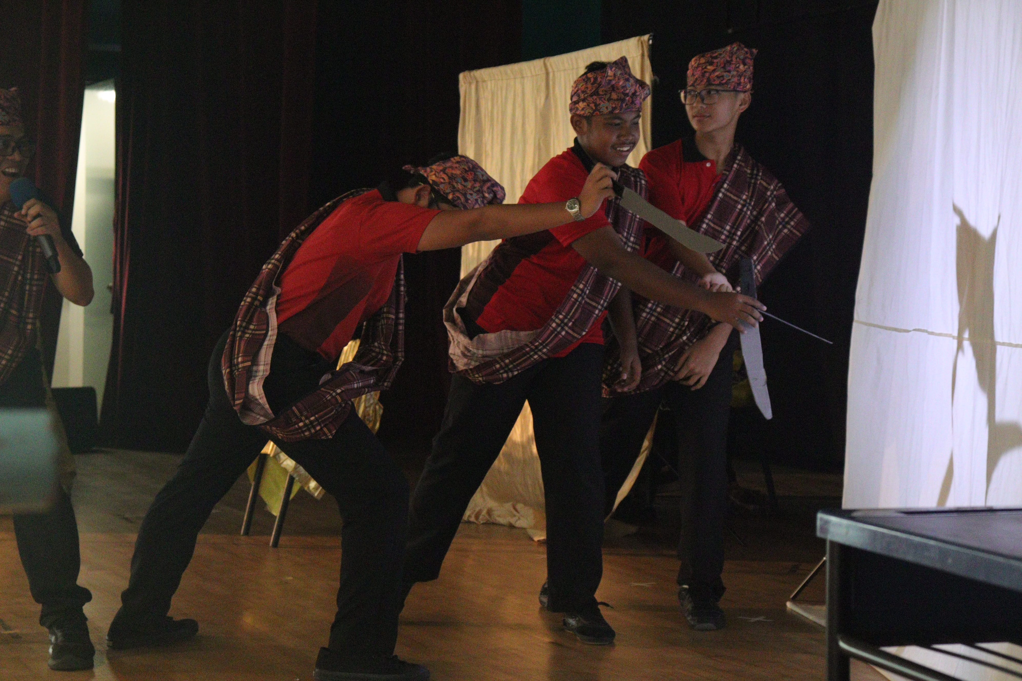Four young performers in red shirts, plaid wraps, and patterned caps perform on a wooden stage.