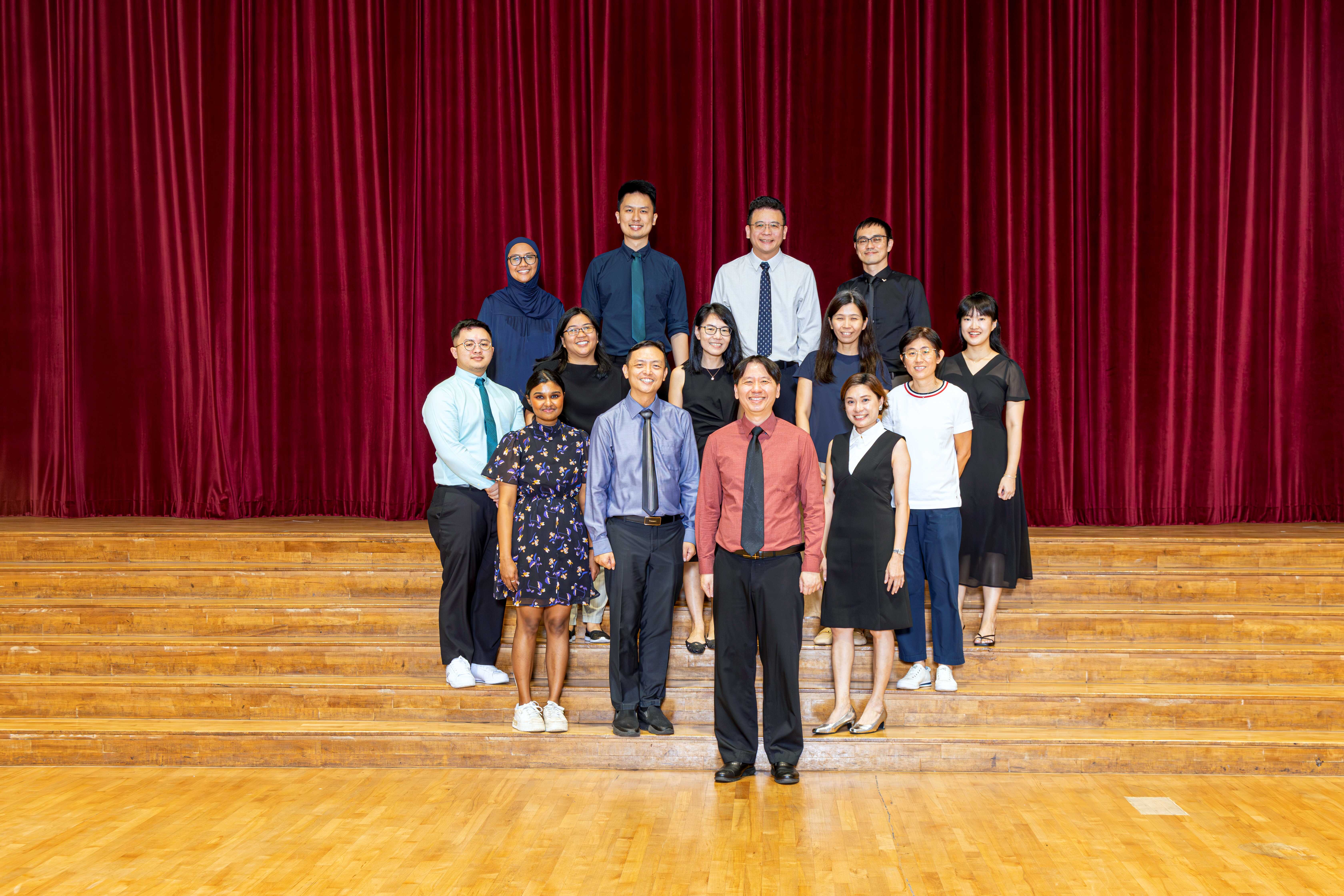 A group of about 13 people posing on wooden stage steps in front of a red curtain.