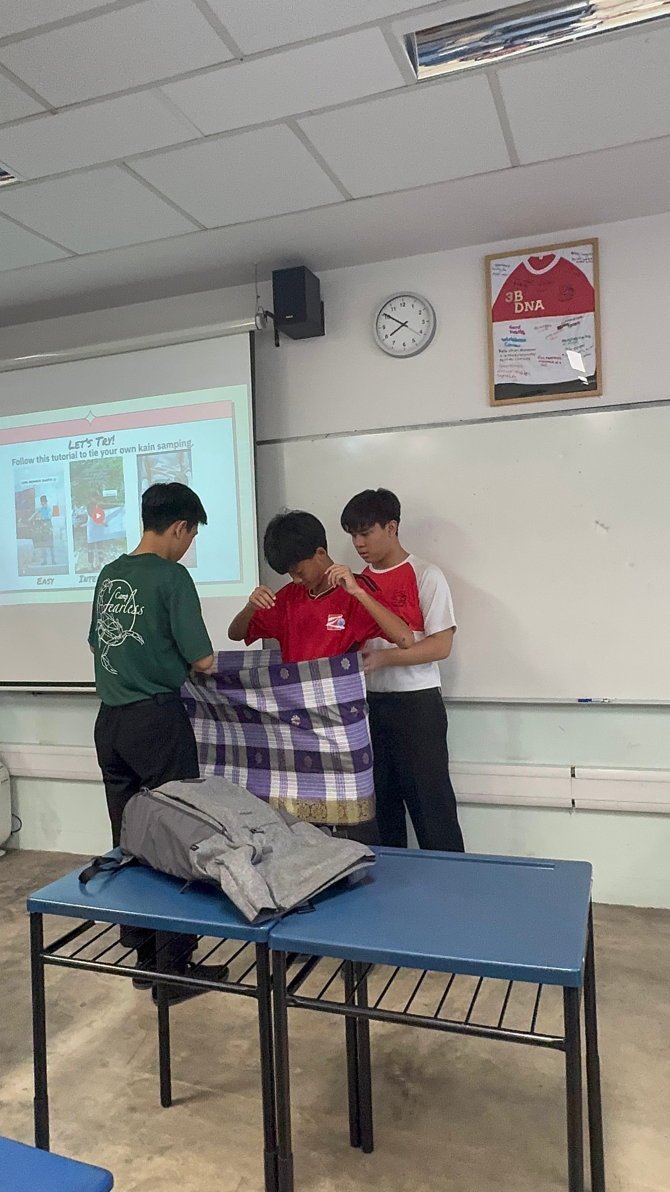Three students in a classroom practice tying a kain samping fabric, with a tutorial projected on screen behind them.