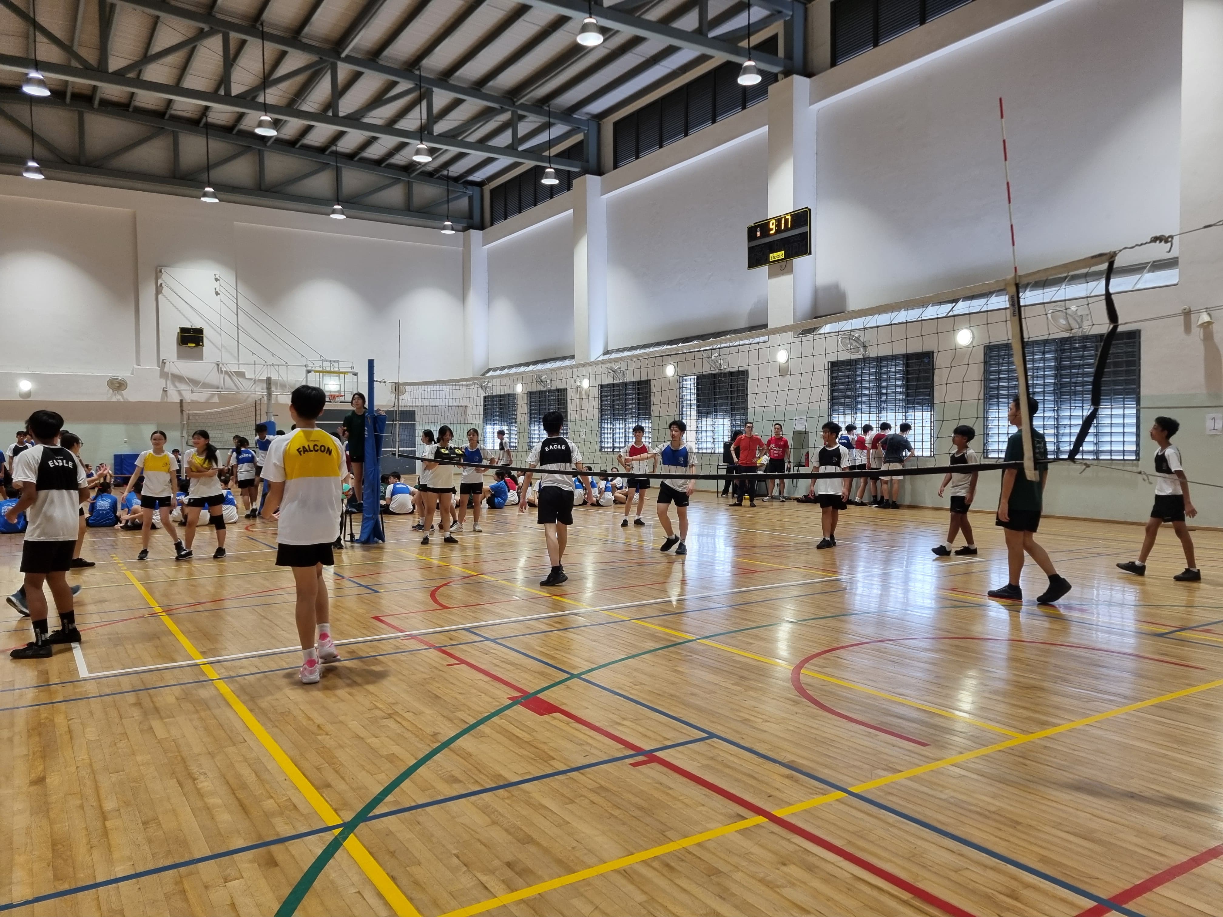 Students in Falcon and Eagle jerseys play volleyball on an indoor court with a scoreboard showing 9-11.