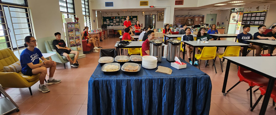 Students gathered in a room with a blue-draped food table holding trays and drink dispensers, seated at yellow and red chairs.