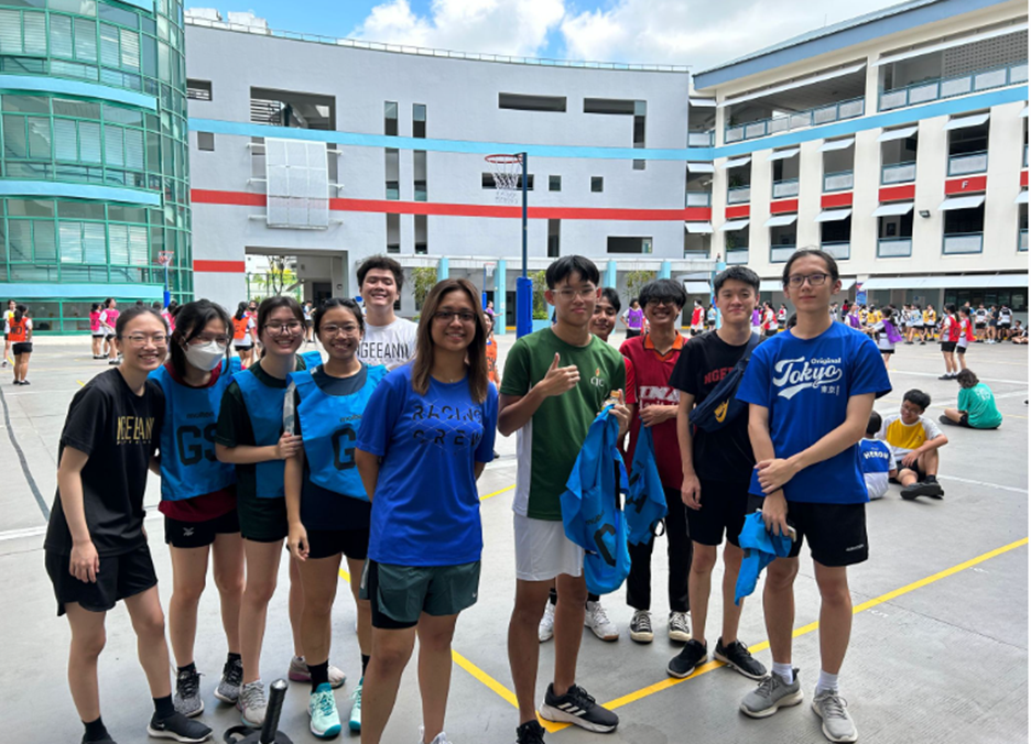 A group of about ten students posing together on a school basketball court outdoors, wearing casual and blue sports bibs.