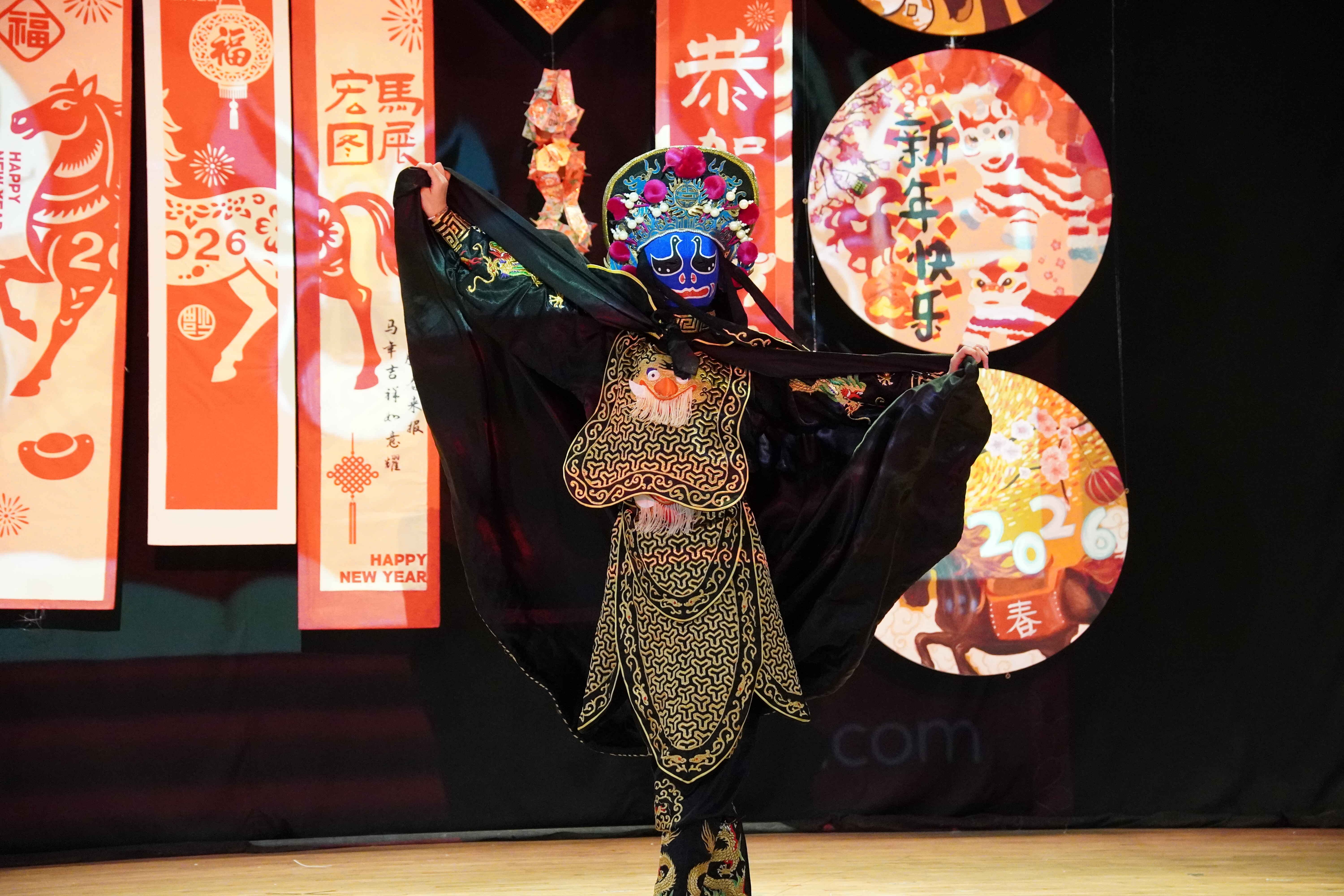 Performer in traditional Chinese opera costume with blue painted mask and black embroidered robe on stage with Lunar New Year decorations.