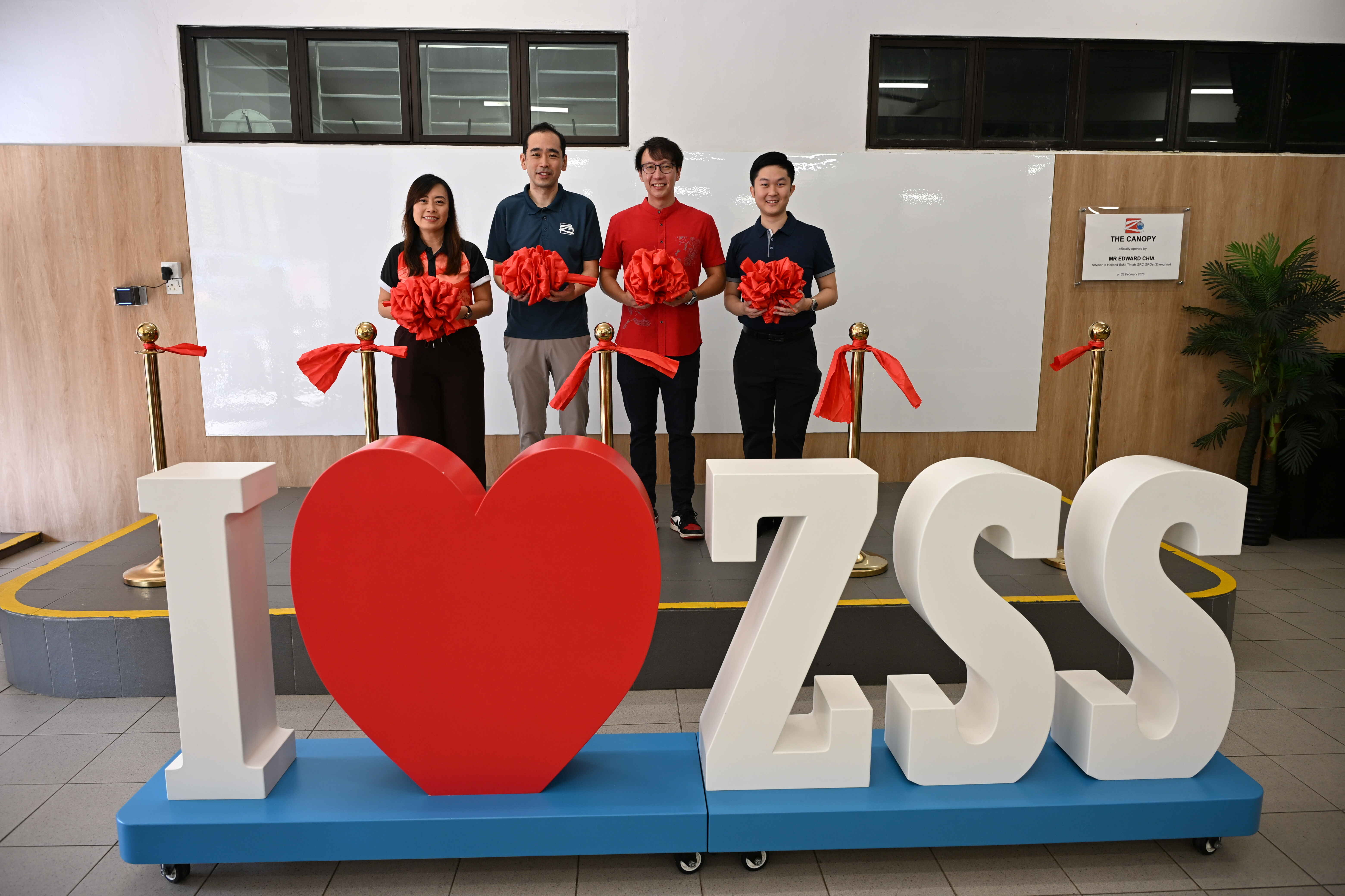 Four people holding red flowers stand behind a large I Love ZSS sign with a red heart at an indoor ceremony.