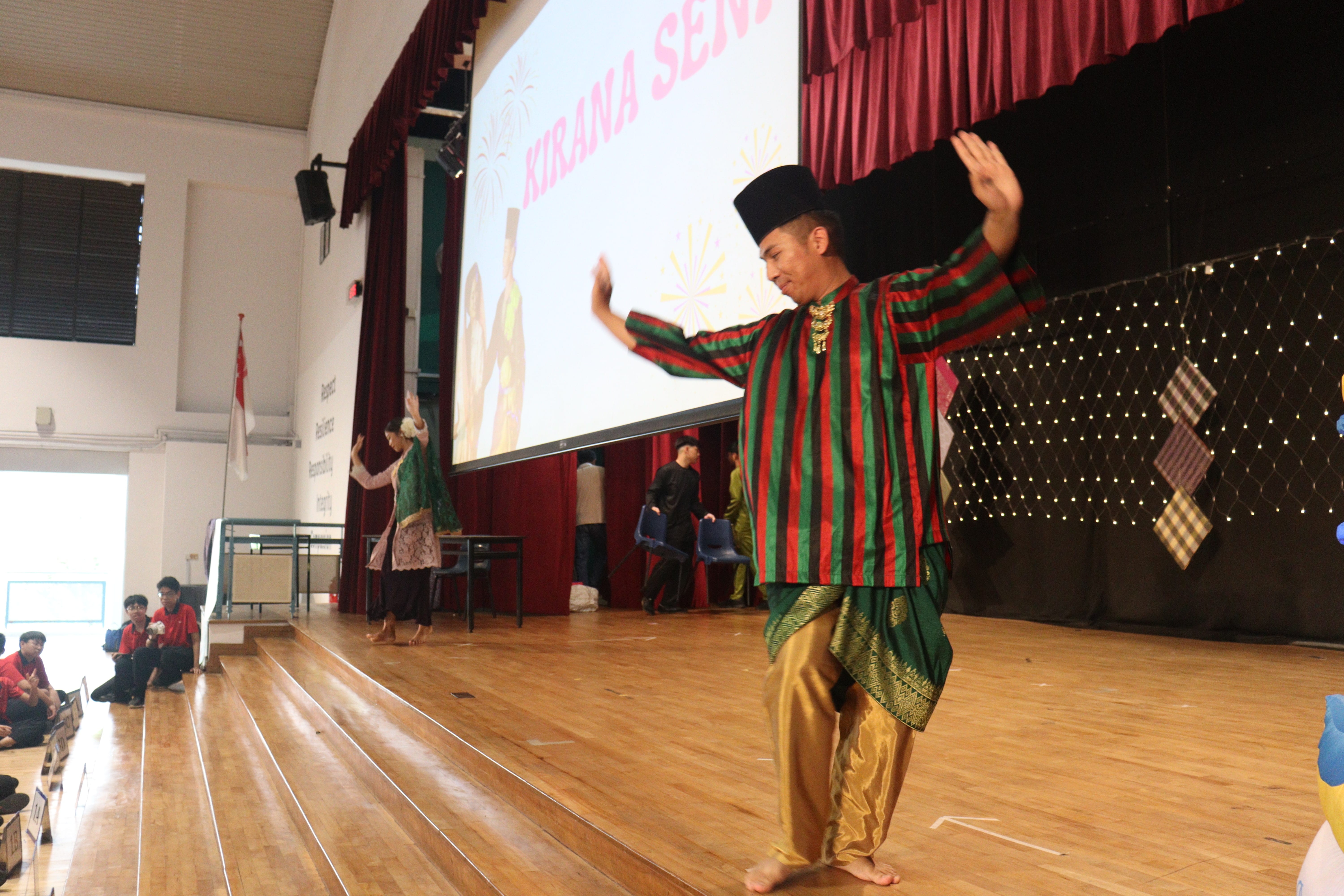 Performers in traditional Malay costumes dance on a school hall stage with a Kirana Seni event screen behind them.