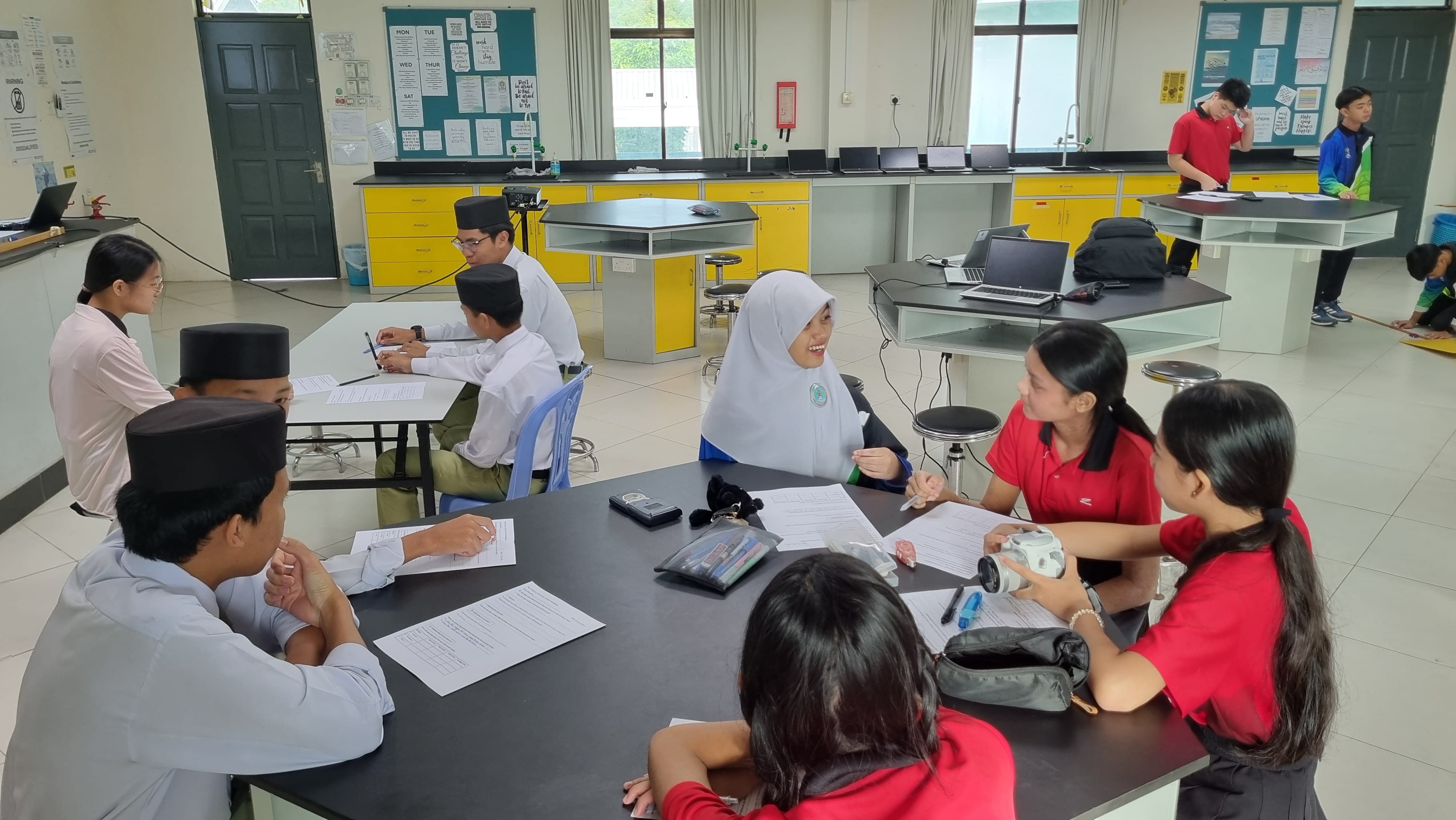 Students in school uniforms sit around tables with papers in a science classroom with yellow cabinets.