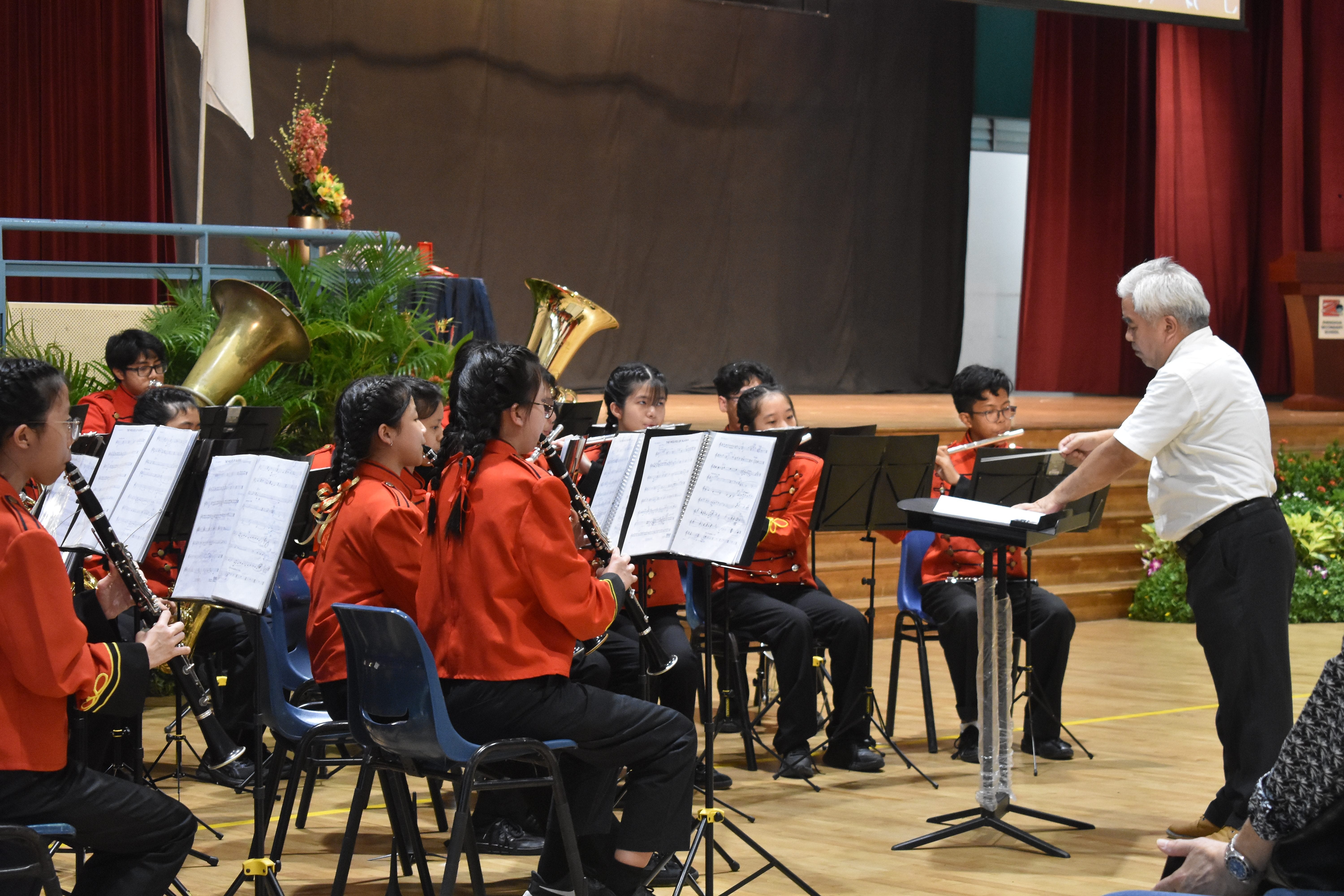 A conductor leads young students in red uniforms playing wind instruments on a school stage with music stands.