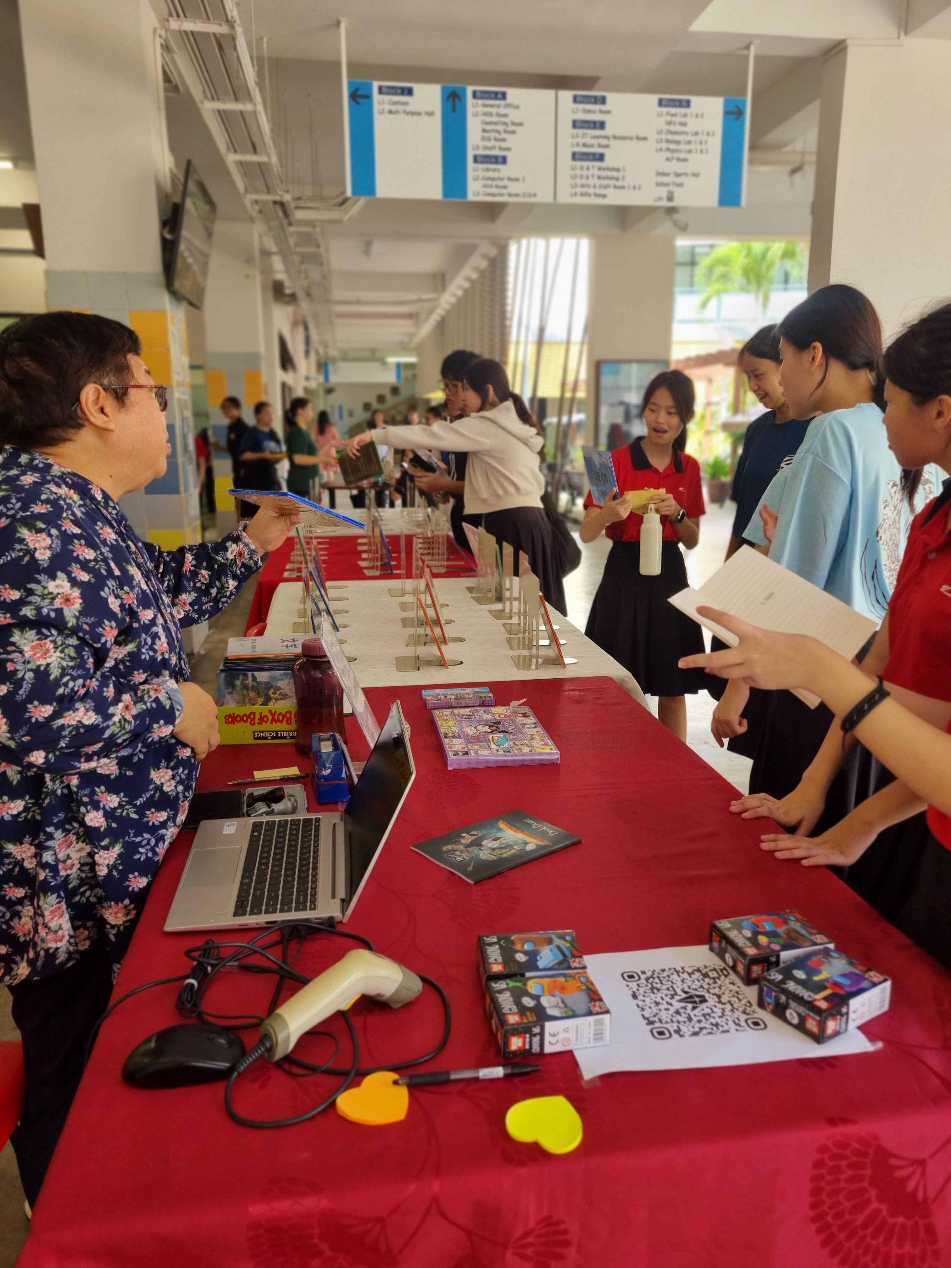 Students browse a red-covered table with books, a laptop, and a barcode scanner at a school event.