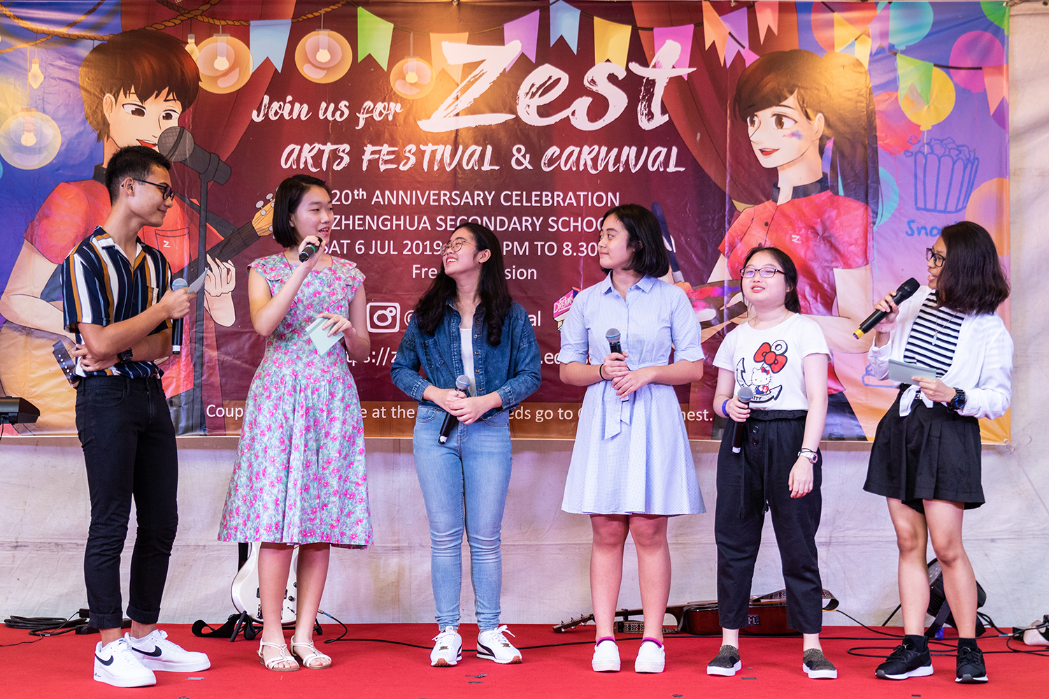 Six young students stand on a red stage holding microphones in front of a Zest Arts Festival banner.