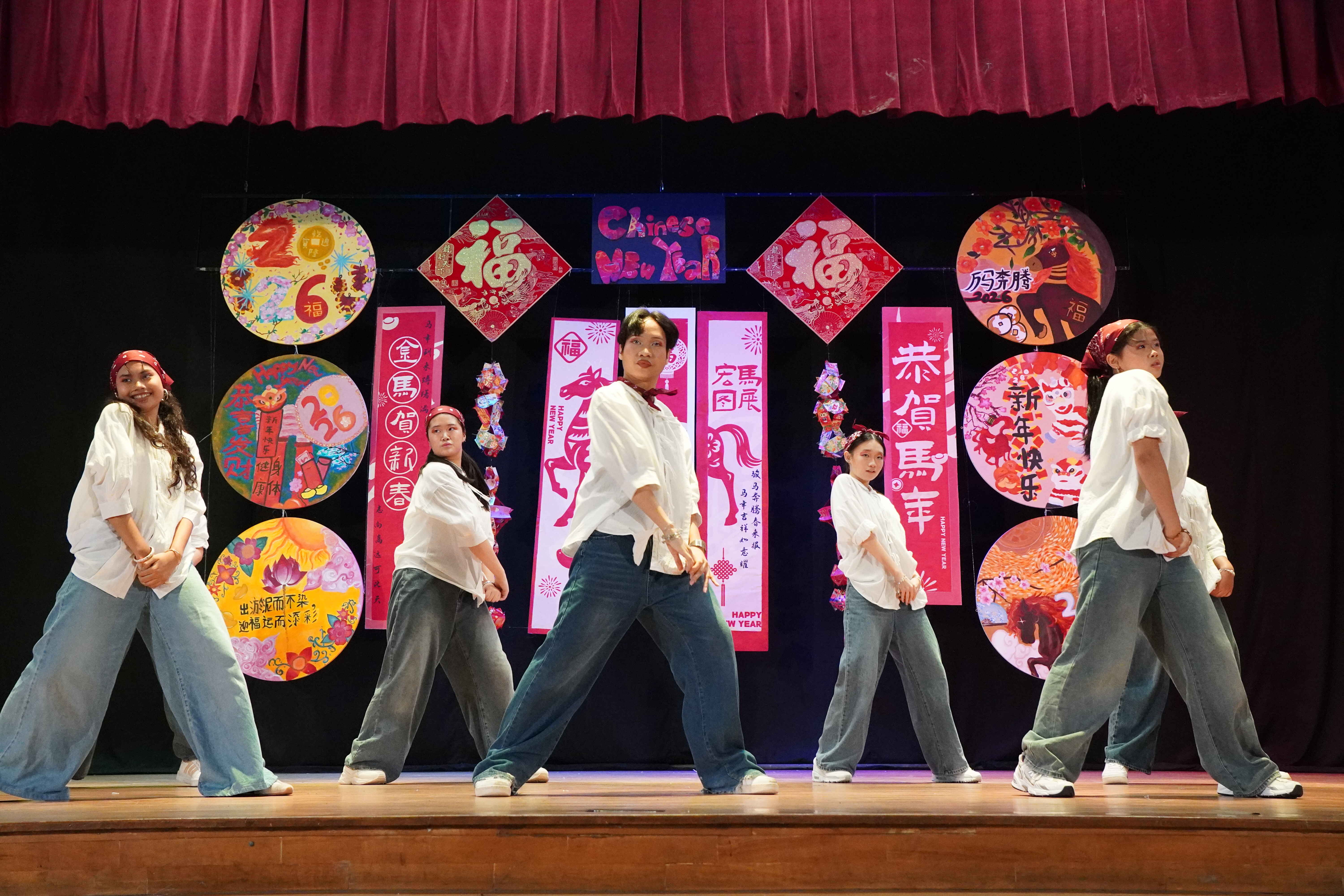 Five performers in white shirts and jeans dance on a stage decorated with colorful Chinese New Year banners and artwork.
