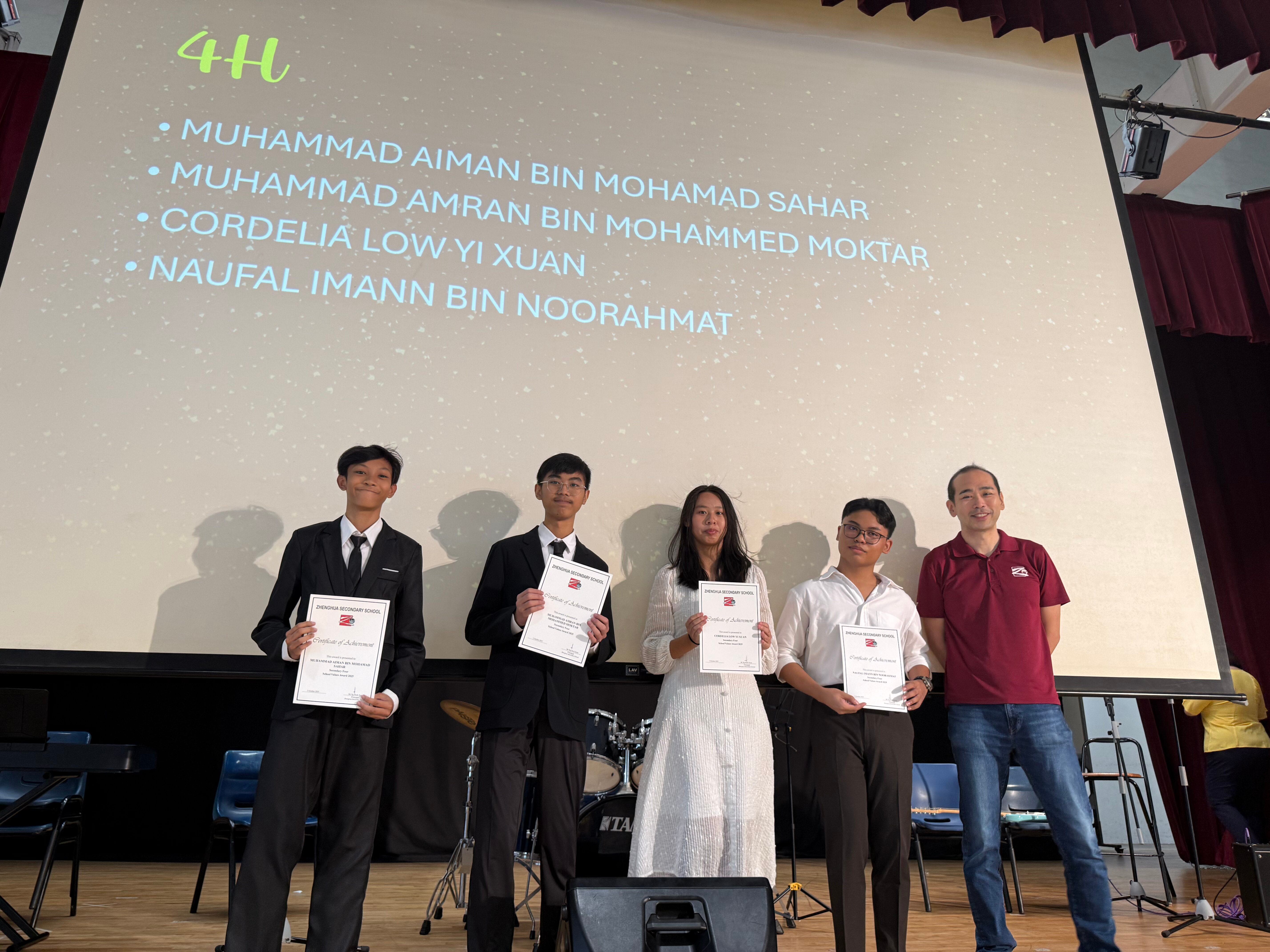 Four students holding certificates stand with a teacher on stage before a screen listing class 4H student names.