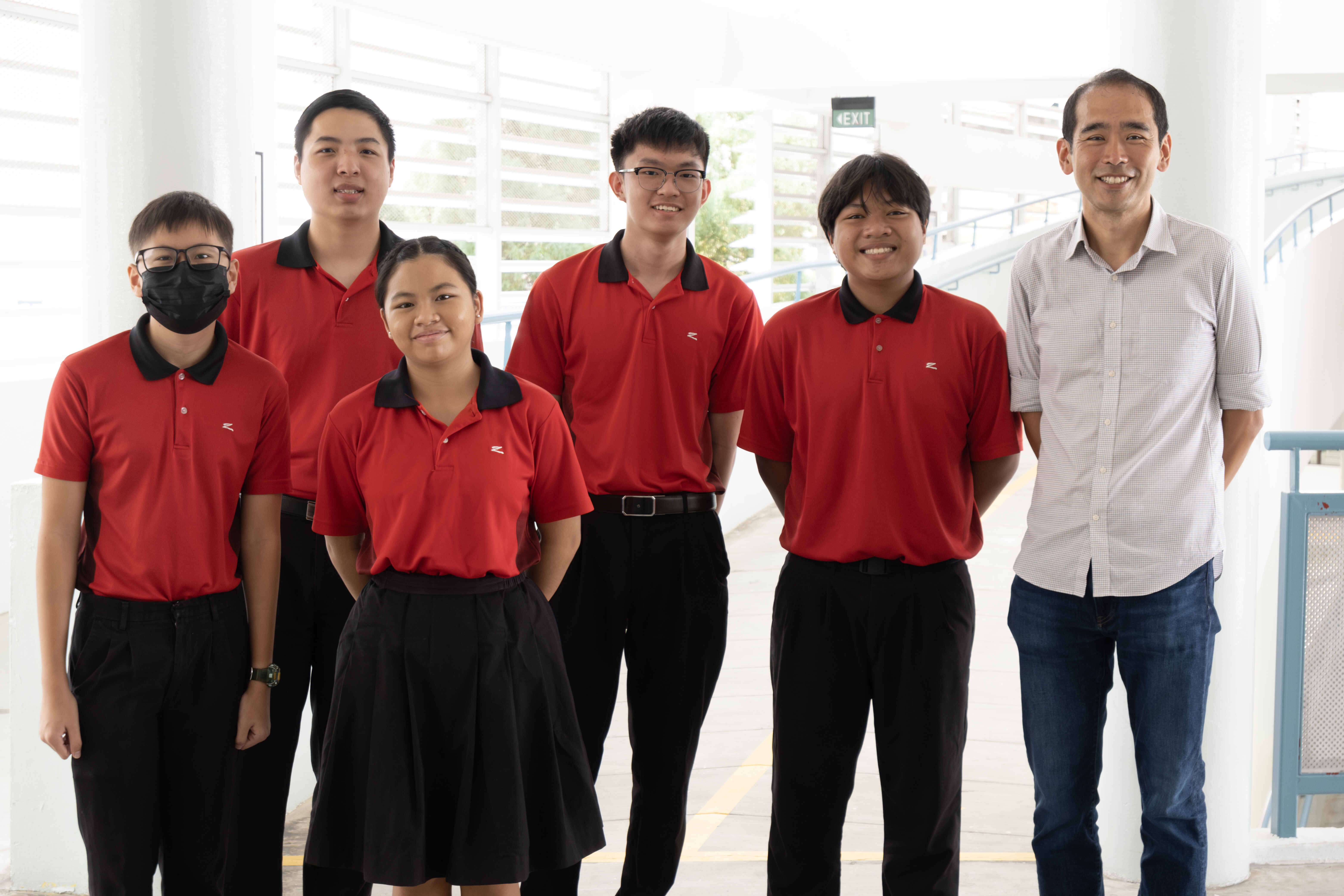 Five students in red and black uniforms pose with a smiling adult man in a gray shirt indoors.