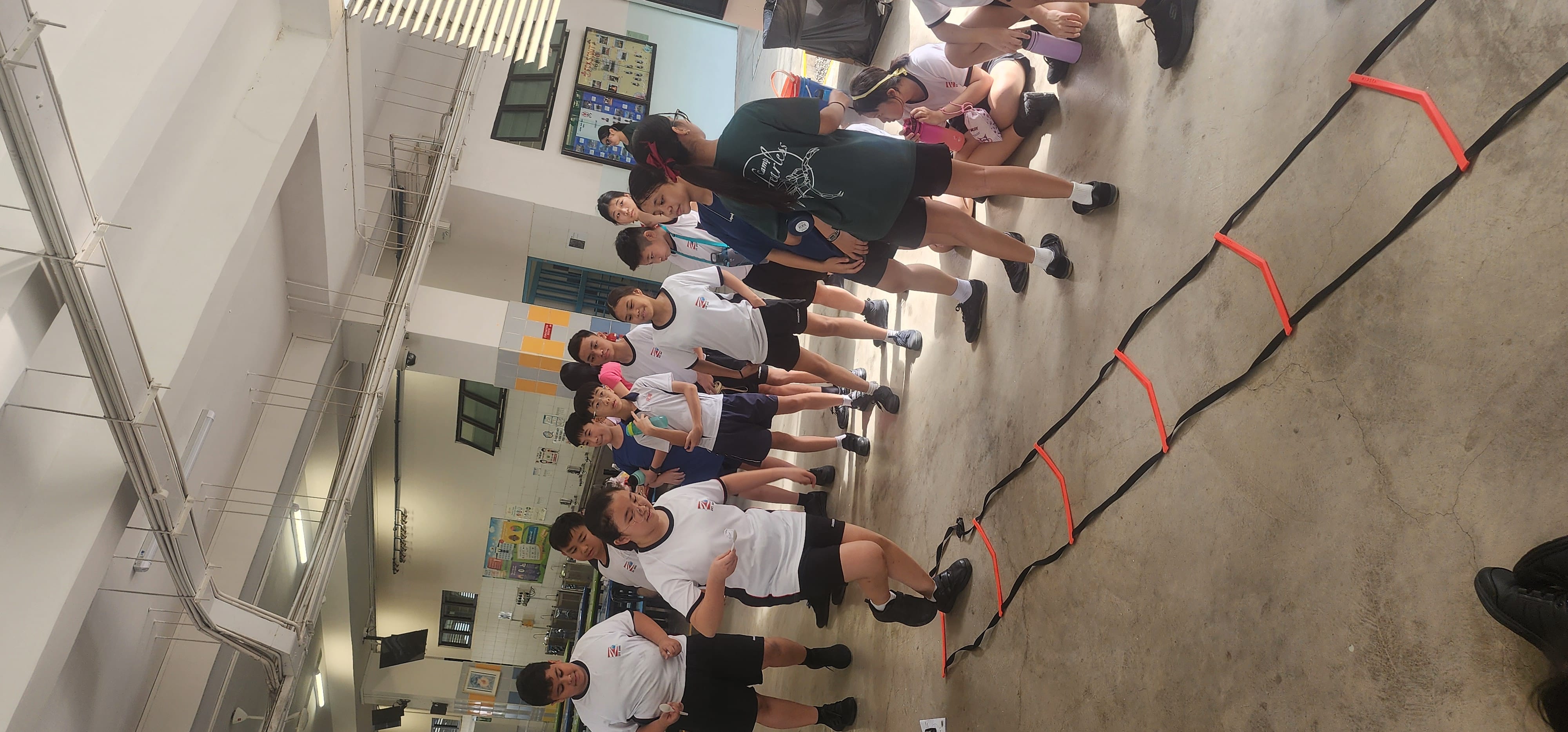 Students in school uniforms stand around an agility ladder on a concrete floor inside a covered hall.