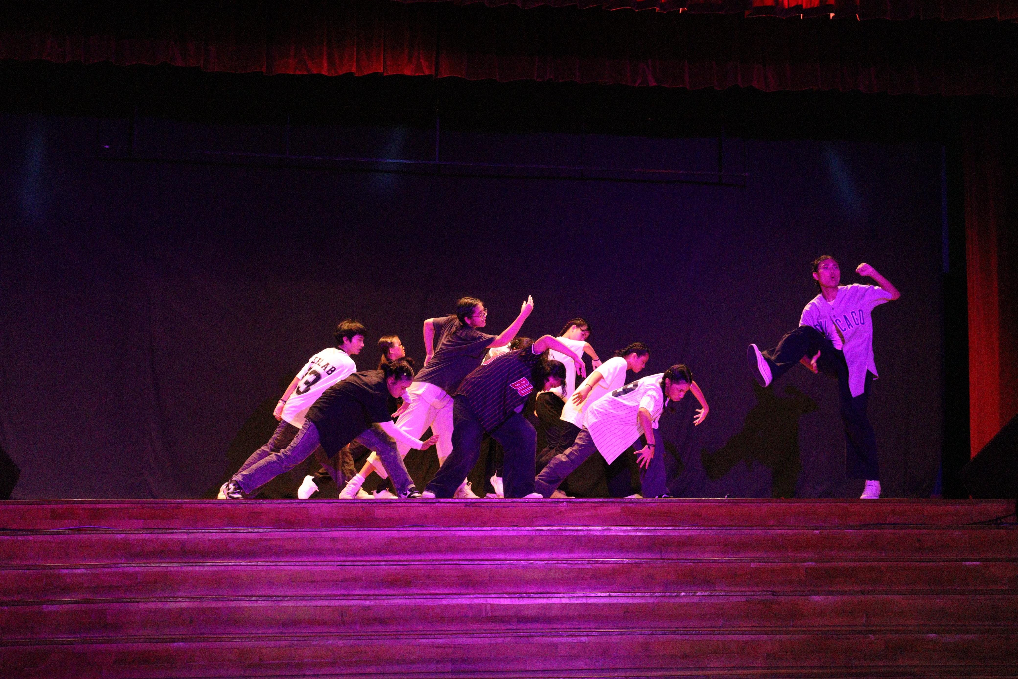 A group of young dancers in white and black outfits performing hip-hop on a dimly lit stage with pink lighting.