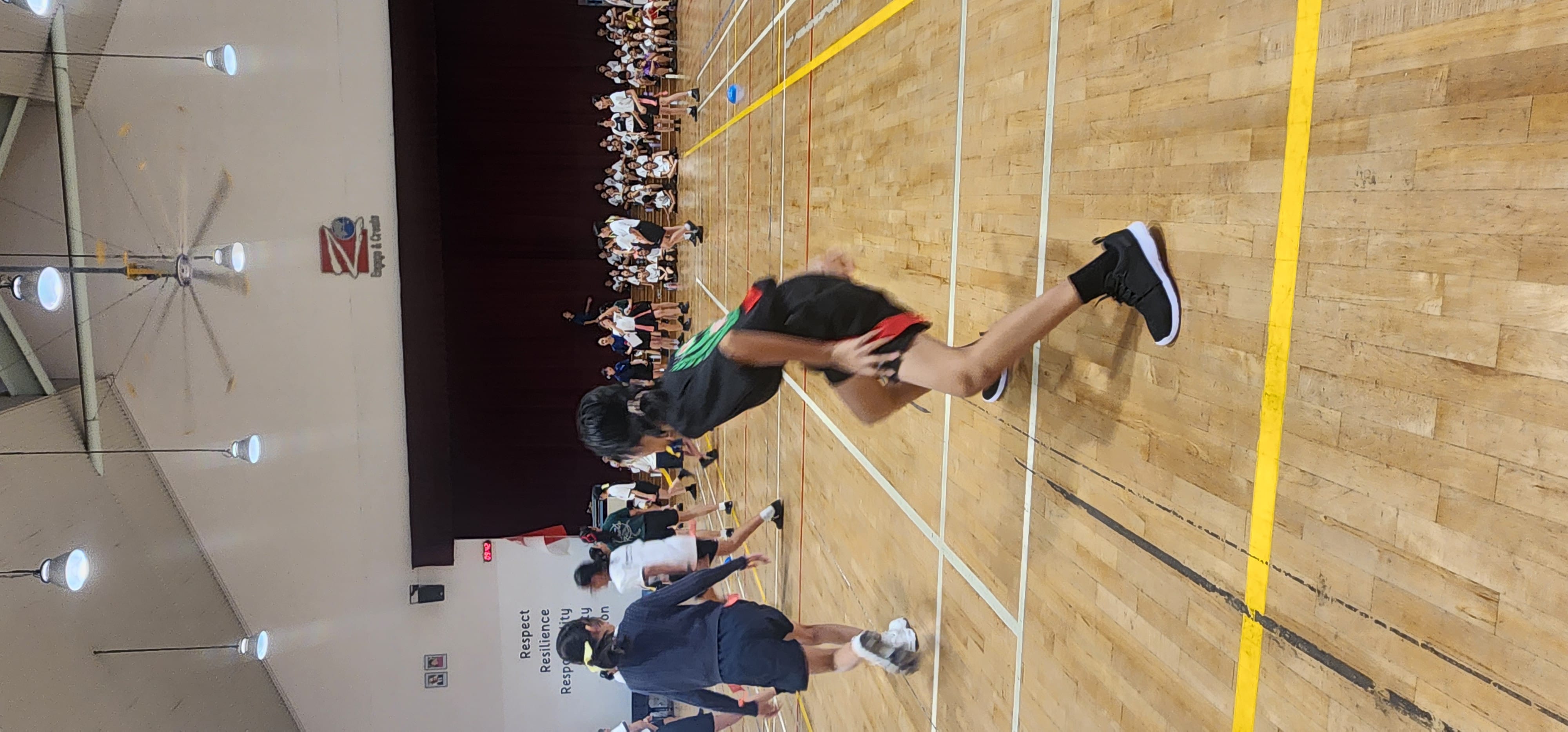 Students run and play in a school gymnasium with wooden floors while others sit watching along the walls.