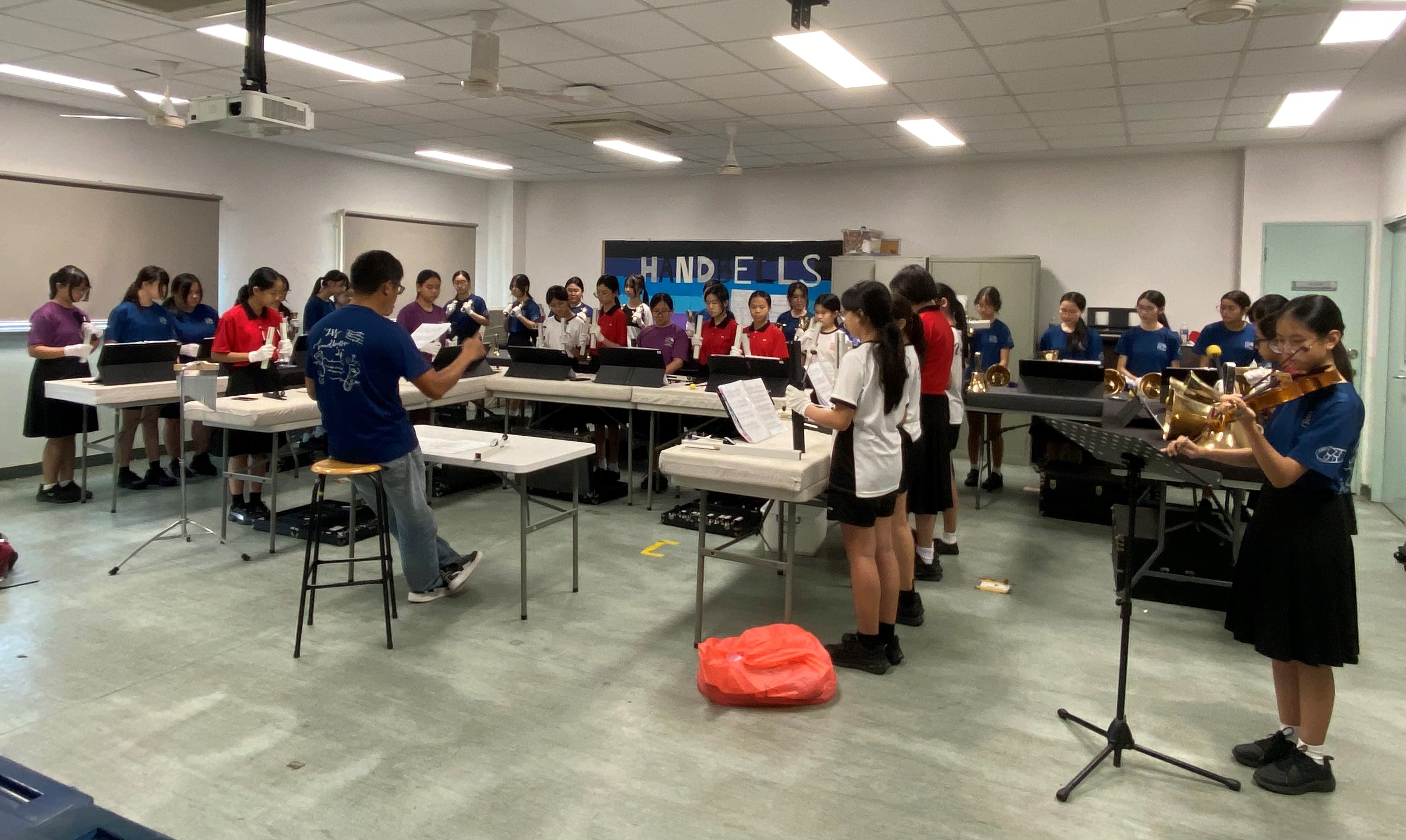 Students rehearse in a music room with a conductor, playing brass instruments and handbells at tables with sheet music.