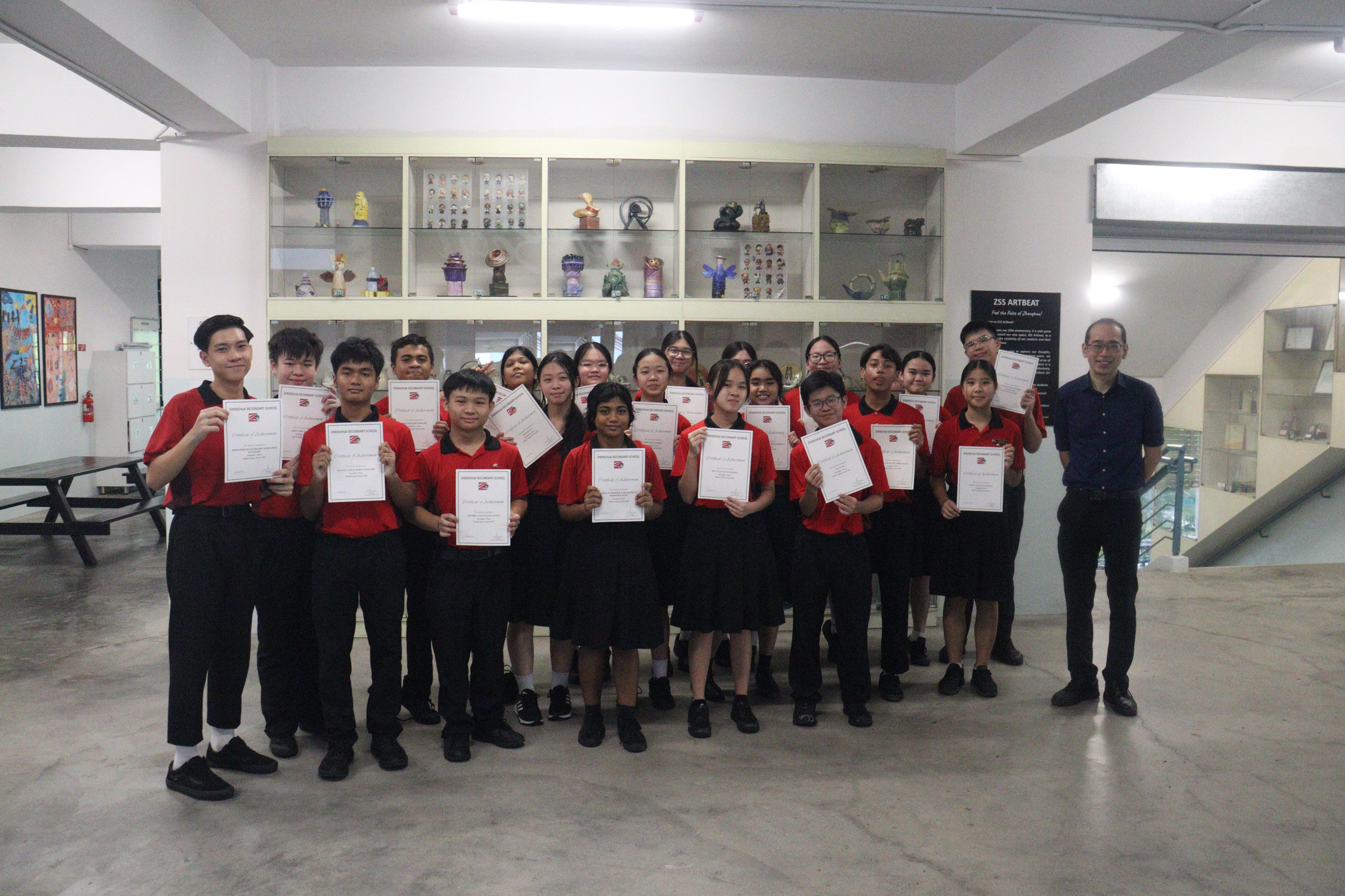 Students in red uniforms hold certificates indoors in front of display shelves with colorful art pieces.