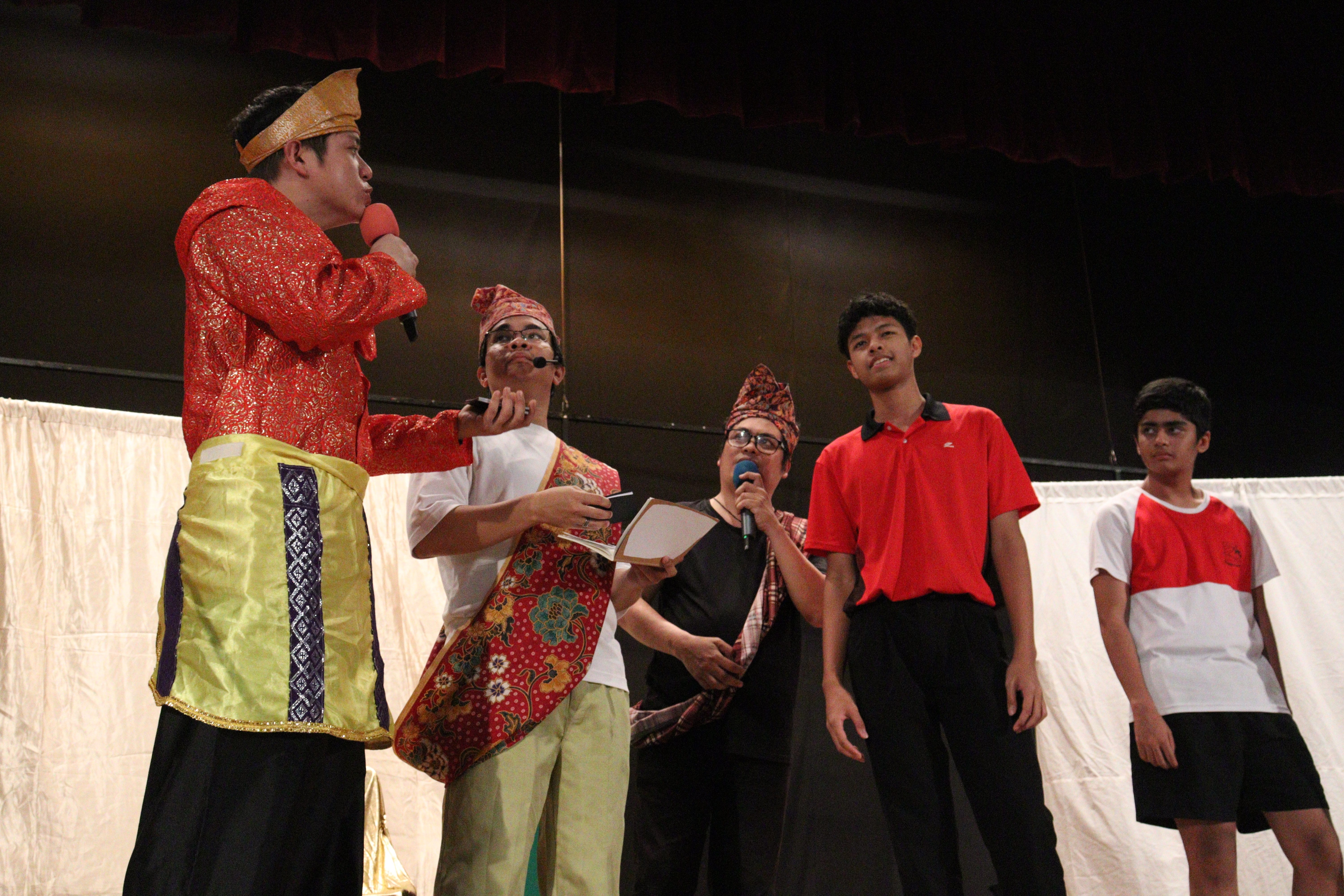 Five young males on stage, some in traditional Southeast Asian costumes with headwear, holding microphones during a performance.