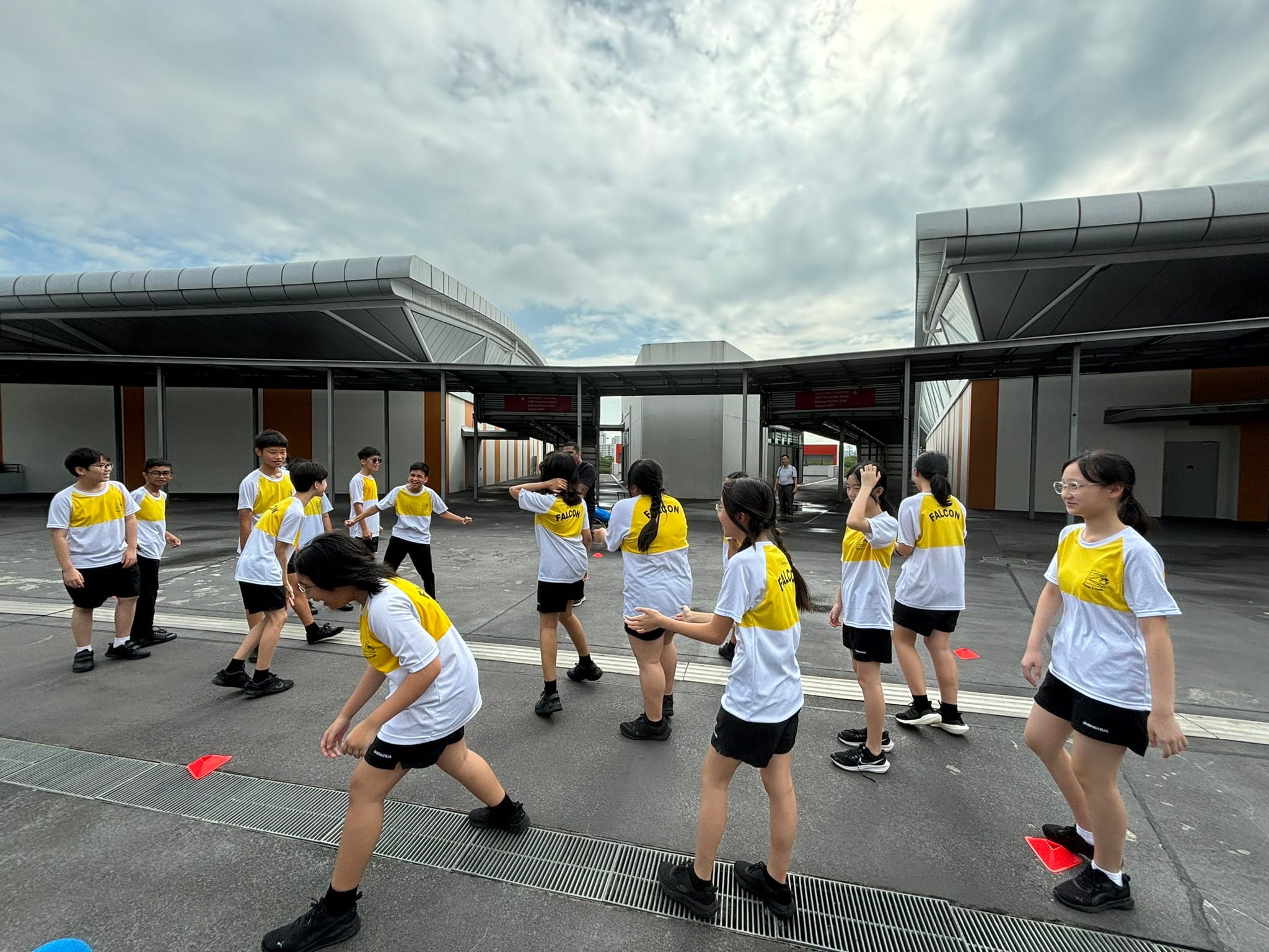 Students in yellow and white Falcon jerseys play an outdoor activity on a school courtyard under a cloudy sky.