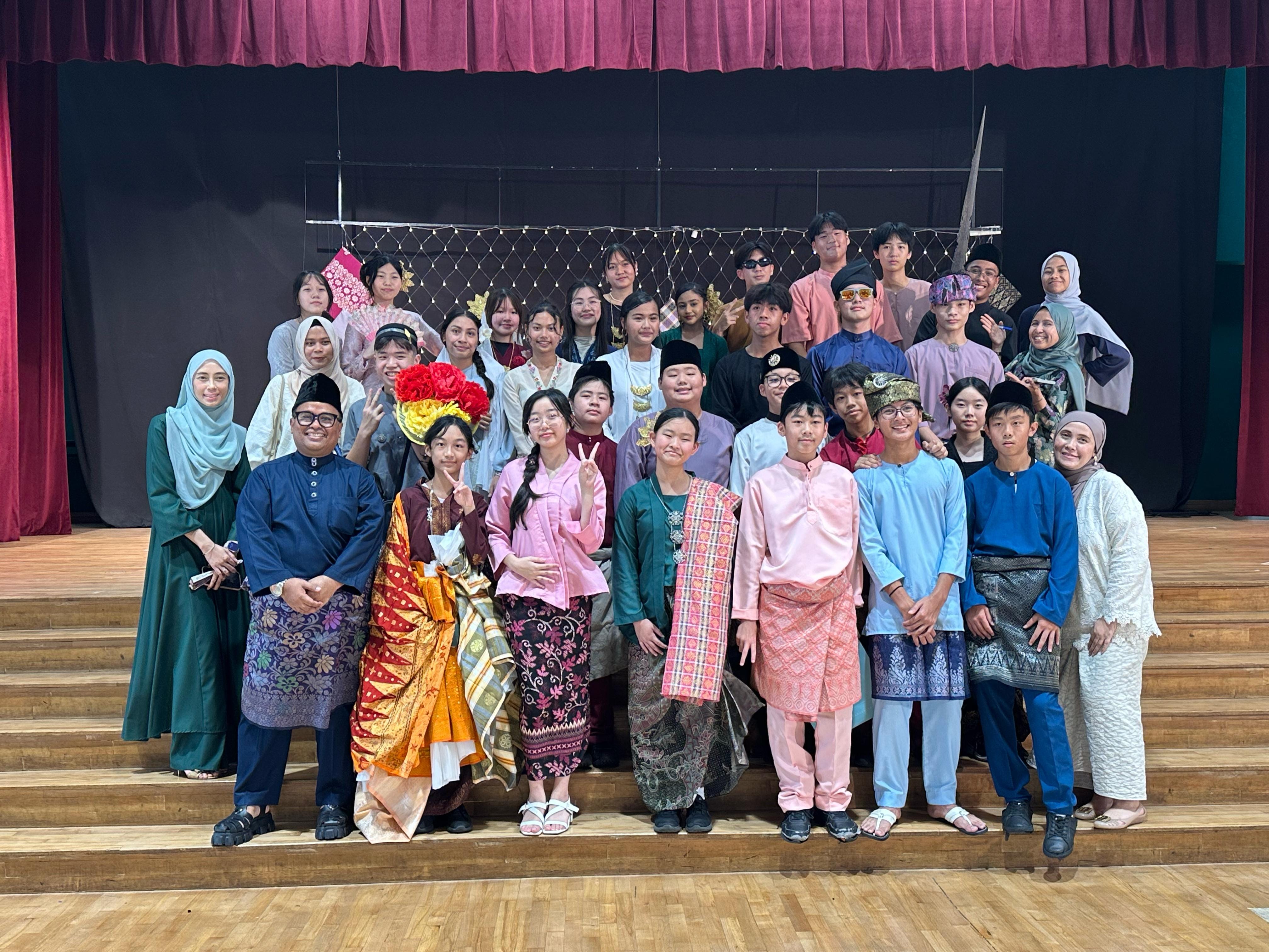 A large group of students and adults in traditional Malay costumes posing on a stage with red curtains.