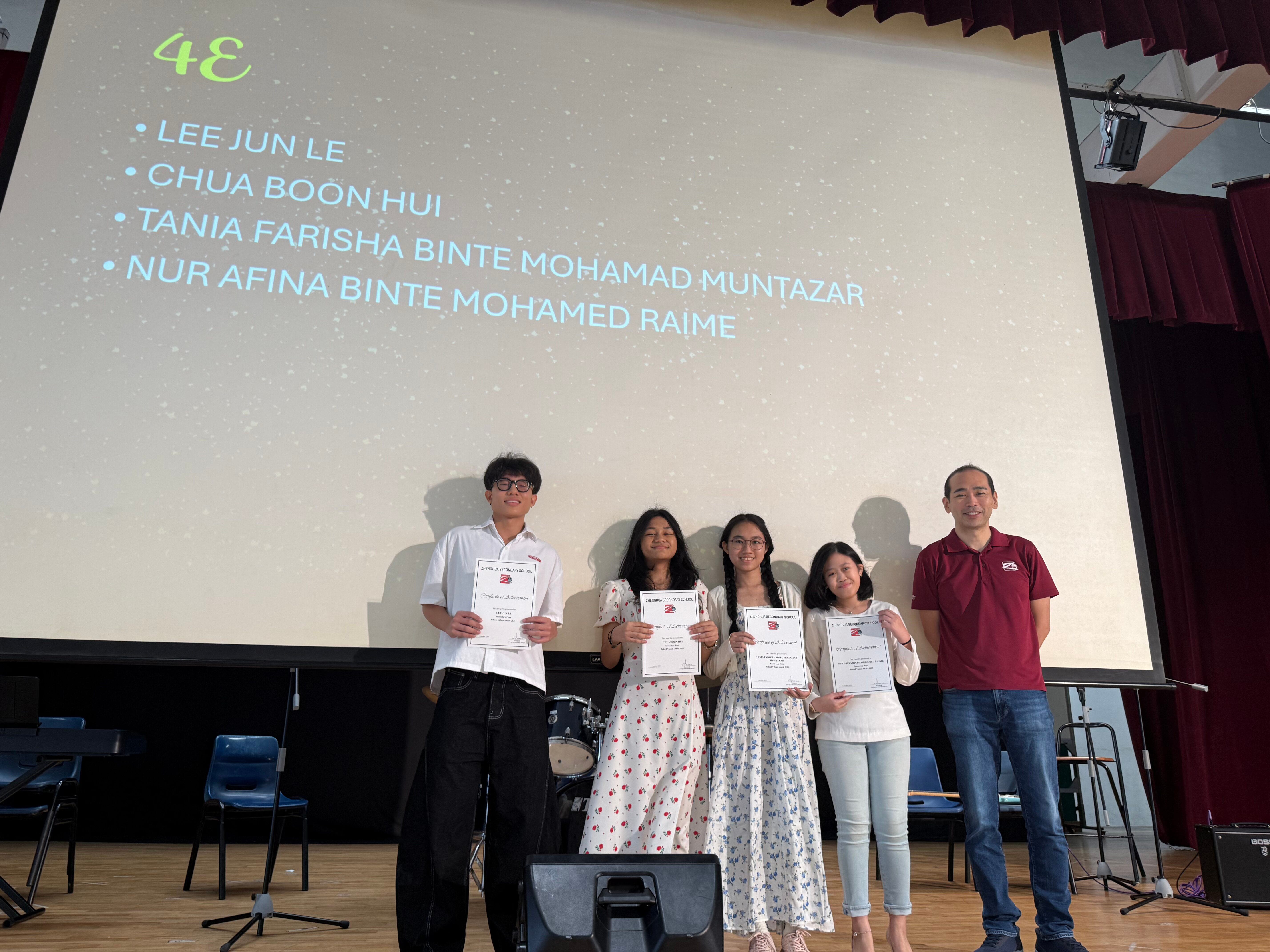 Four students holding certificates stand on stage with a teacher before a projected slide listing class 4E student names.
