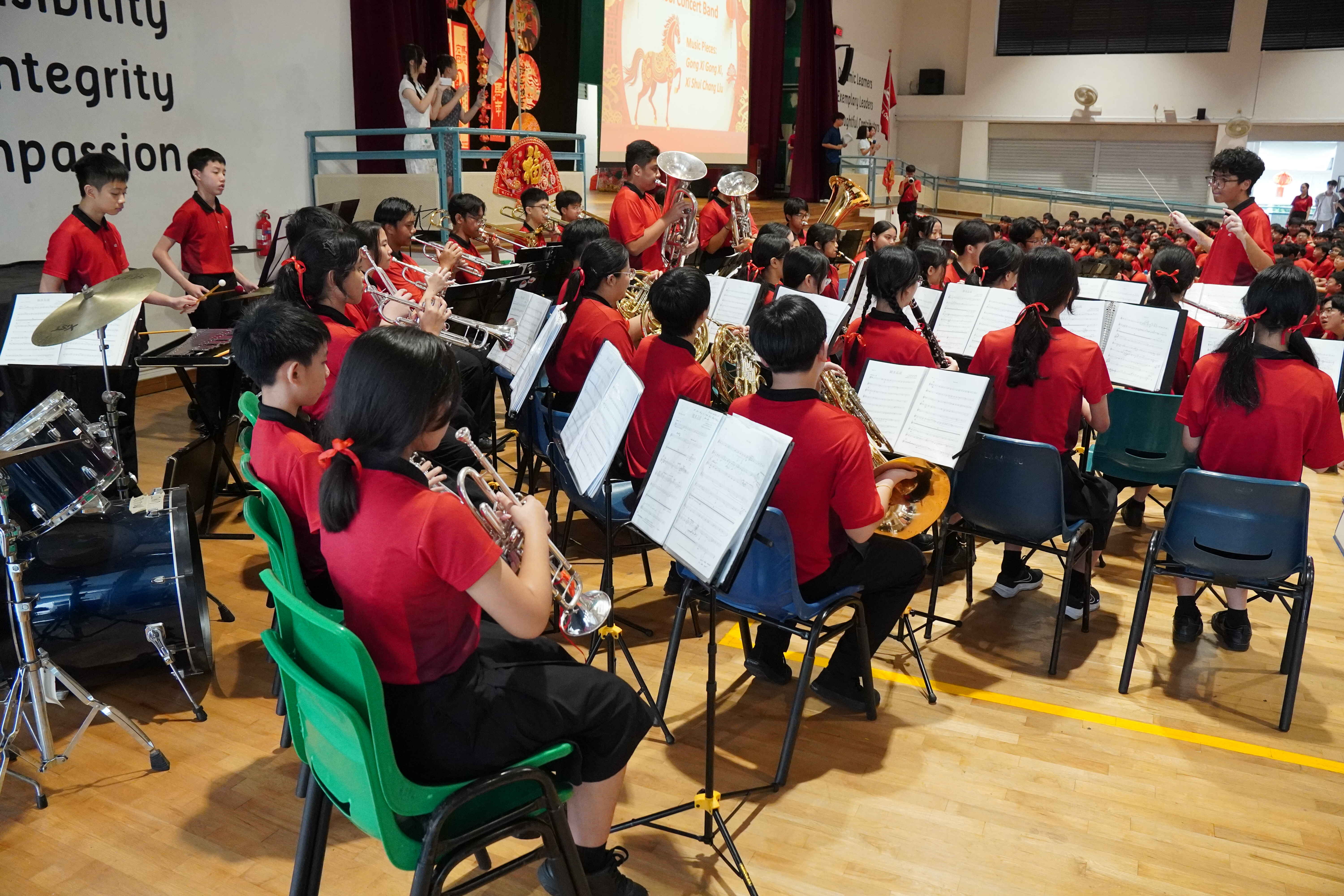 Students in red uniforms perform as a school concert band with brass instruments and drums in a gymnasium.