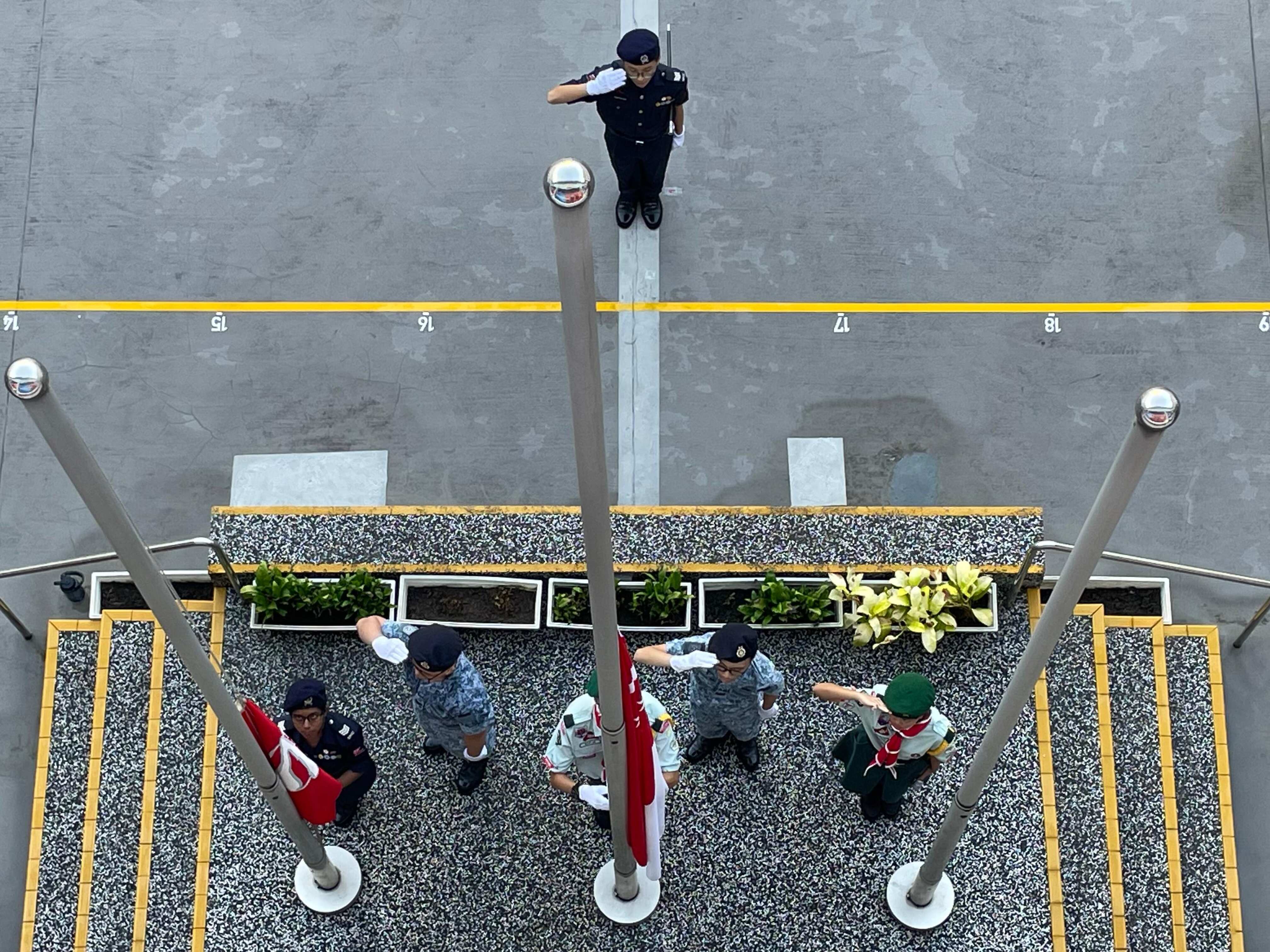 Overhead view of uniformed youth cadets raising flags at flagpoles while one salutes nearby.